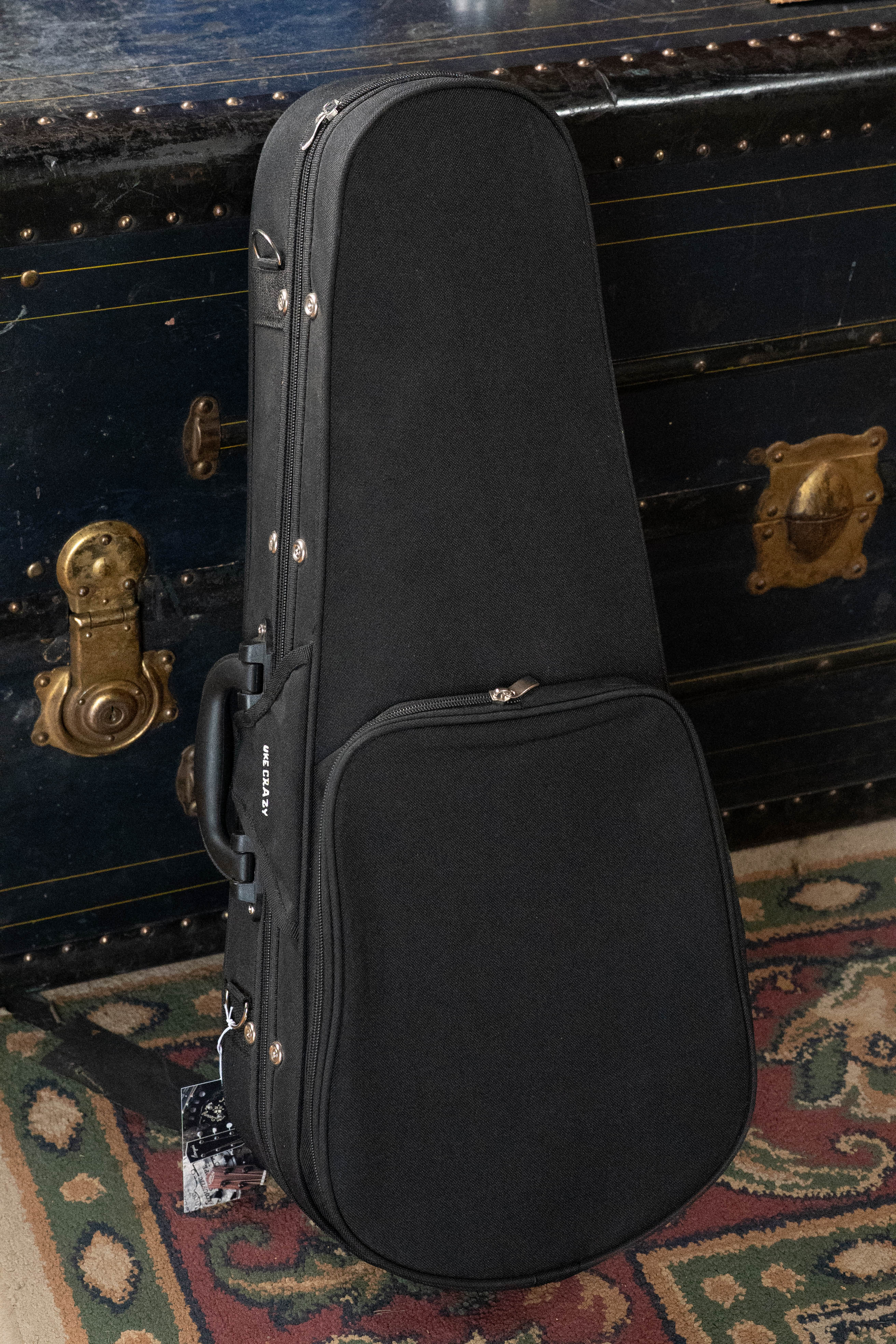 The Kala UC-C Foam Concert Ukulele Hardcase by Kala stands upright on a patterned rug in front of a large antique trunk with brass locks and accents.