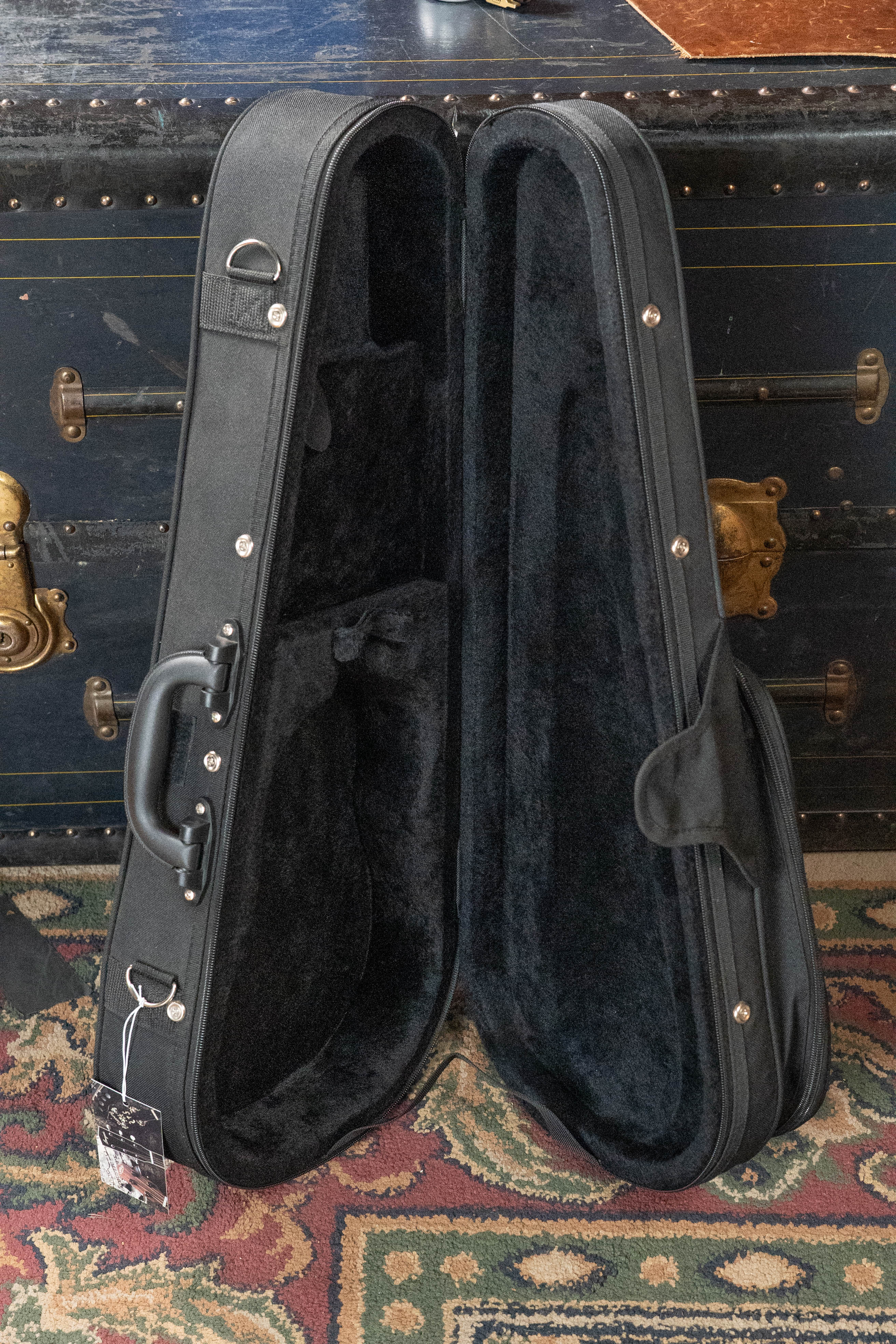 The Kala UC-C Foam Concert Ukulele Hardcase by Kala, black with a velvet-lined interior, stands upright on a patterned carpet in front of a vintage trunk with brass details.