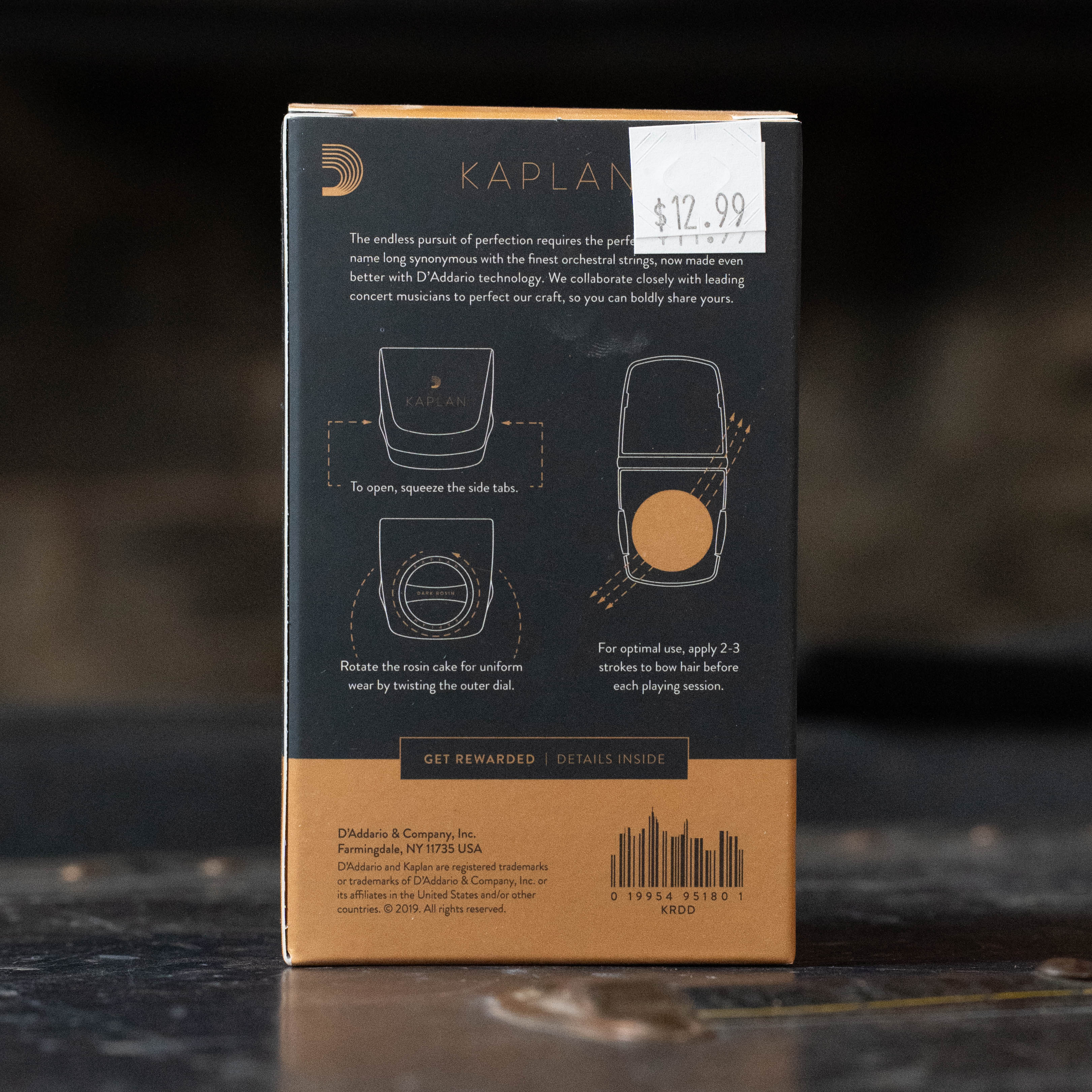 A D'Addario Kaplan Premium Rosin with Case (KRD) box features gold and white text on a dark background, low dust instructions for the included rosin cake, and a $12.99 price sticker on the top right corner. Brand: Kaplan.