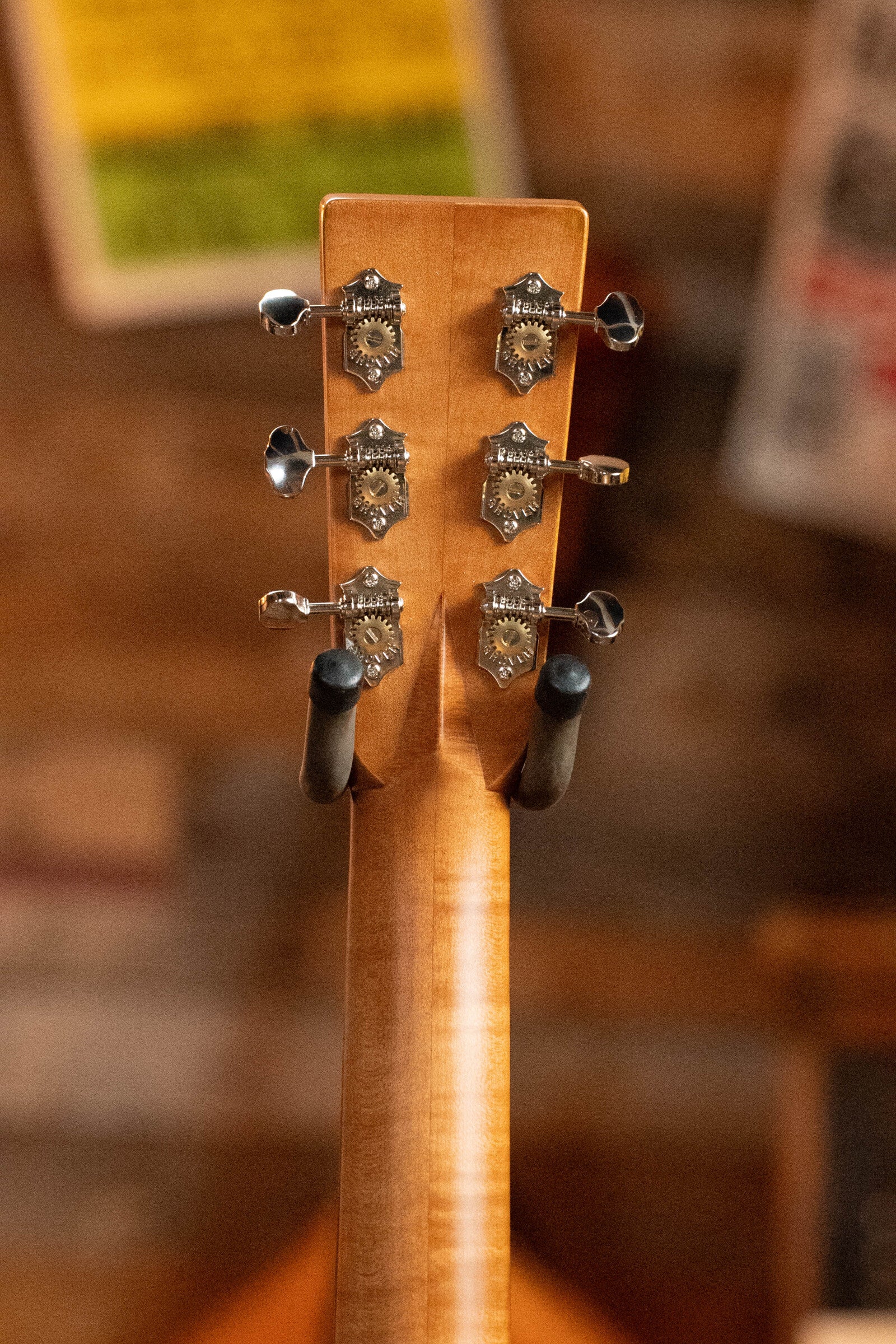 Close-up of the *Limited Edition* Martin & Co. CEO-11 European Spruce/White Oak New Horizons Blue Burst Grand Auditorium Guitar #1204 headstock, showing six tuning pegs and decorative metal gears on a wall hanger against a blurred background.