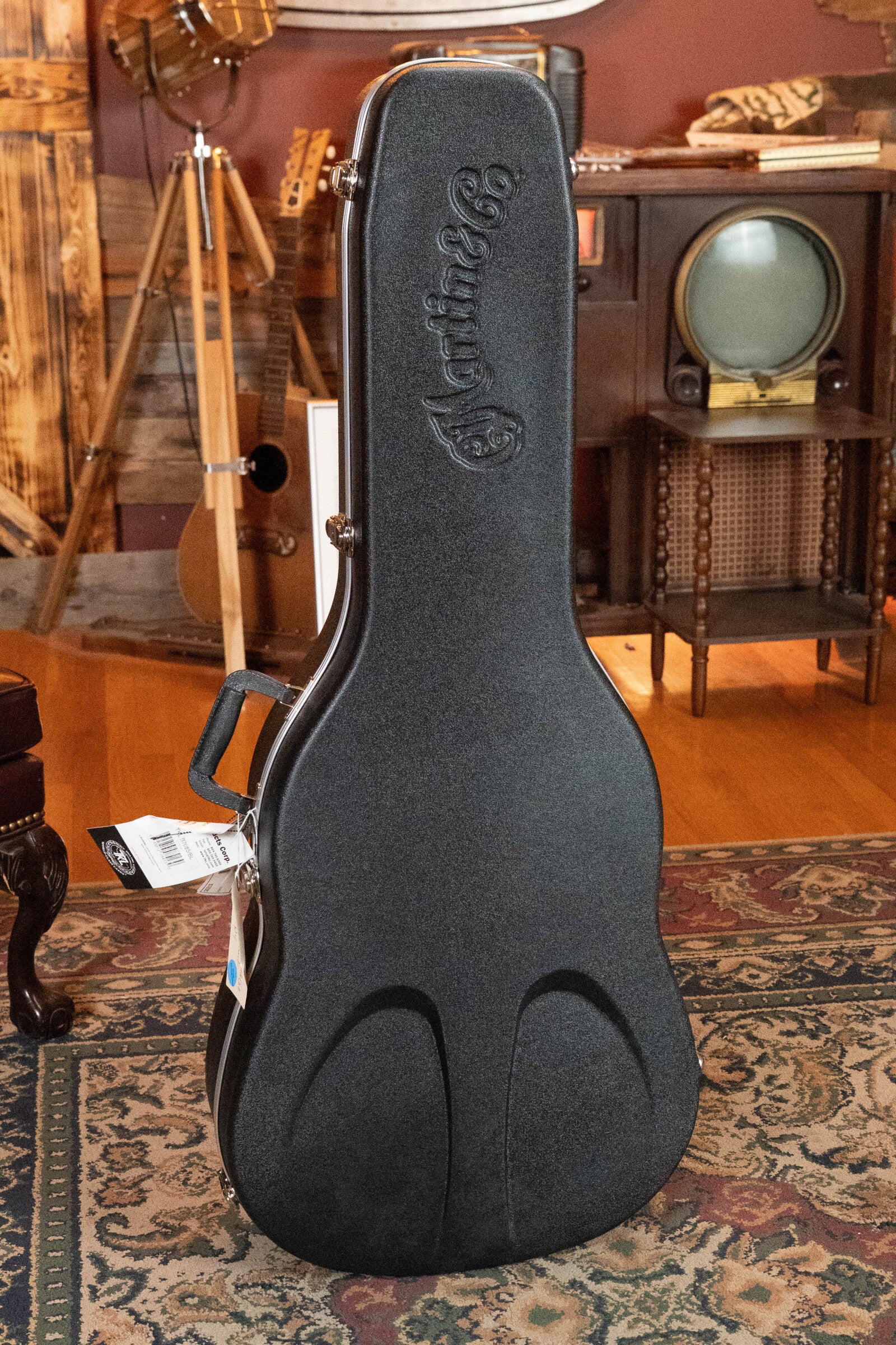 A black Martin & Co. case, possibly holding a *Limited Edition* Martin CEO-11 European Spruce/White Oak New Horizons Blue Burst Grand Auditorium Guitar w/Electronics (#1204), stands upright on a rug in a vintage room with retro TV and wood decor.