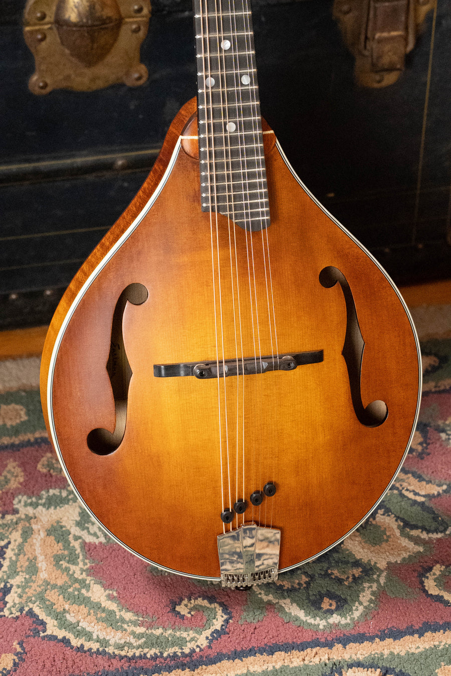A PRE-ORDER DEPOSIT ONLY *New 2026 Model* Eastman MD305 Wide Nut Adirondack/Maple A-Style Mandolin by Eastman Guitars rests on a patterned rug, with a dark vintage trunk in the background.