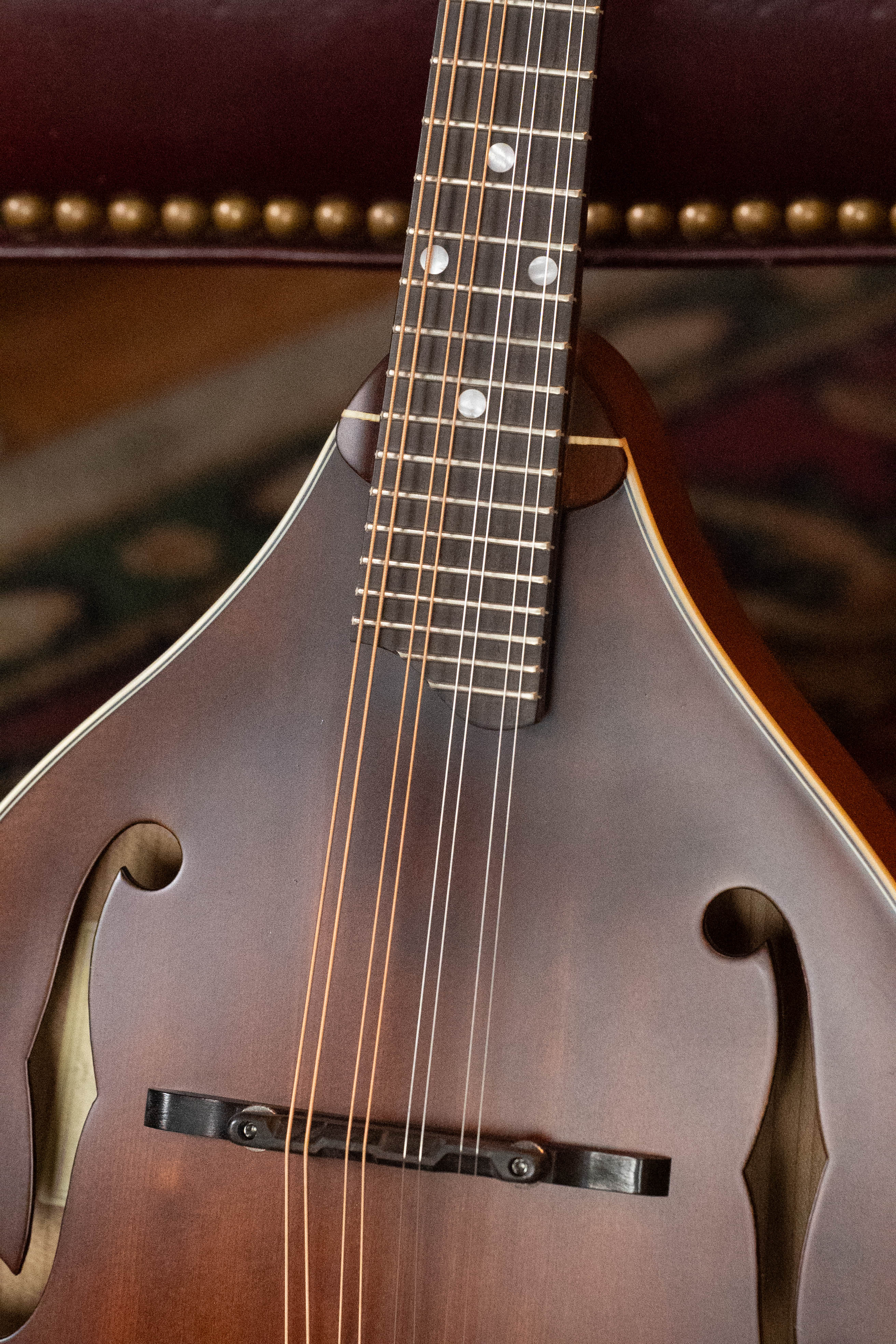 A close-up of the Eastman MD305 Spruce/Maple A-Style Hand-Carved Mandolin #3317 by Eastman Guitars, highlighting its strings, f-holes, and neck with a blurred background of a leather chair and patterned carpet.