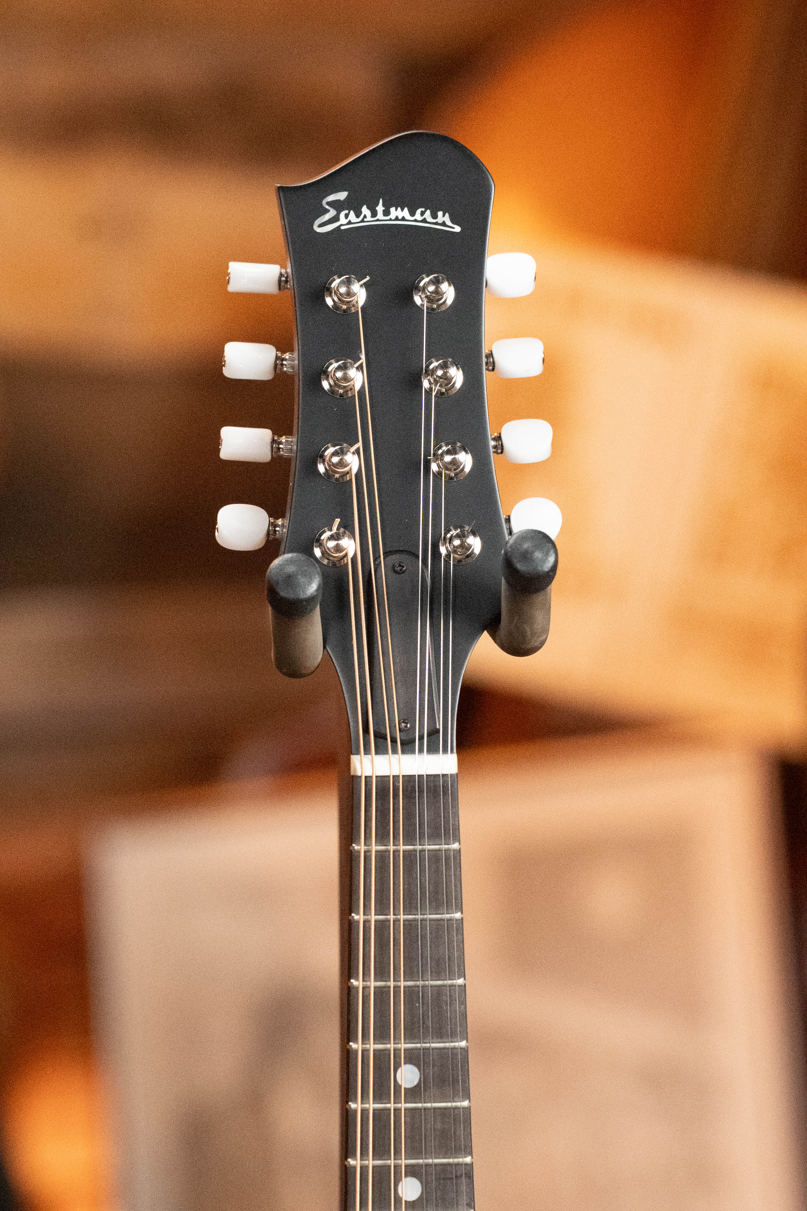 Close-up of the Eastman Guitars MD305 Spruce/Maple A-Style Hand-Carved Mandolin #3317 headstock, featuring white tuning pegs and visible strings against a blurred warm-toned background.
