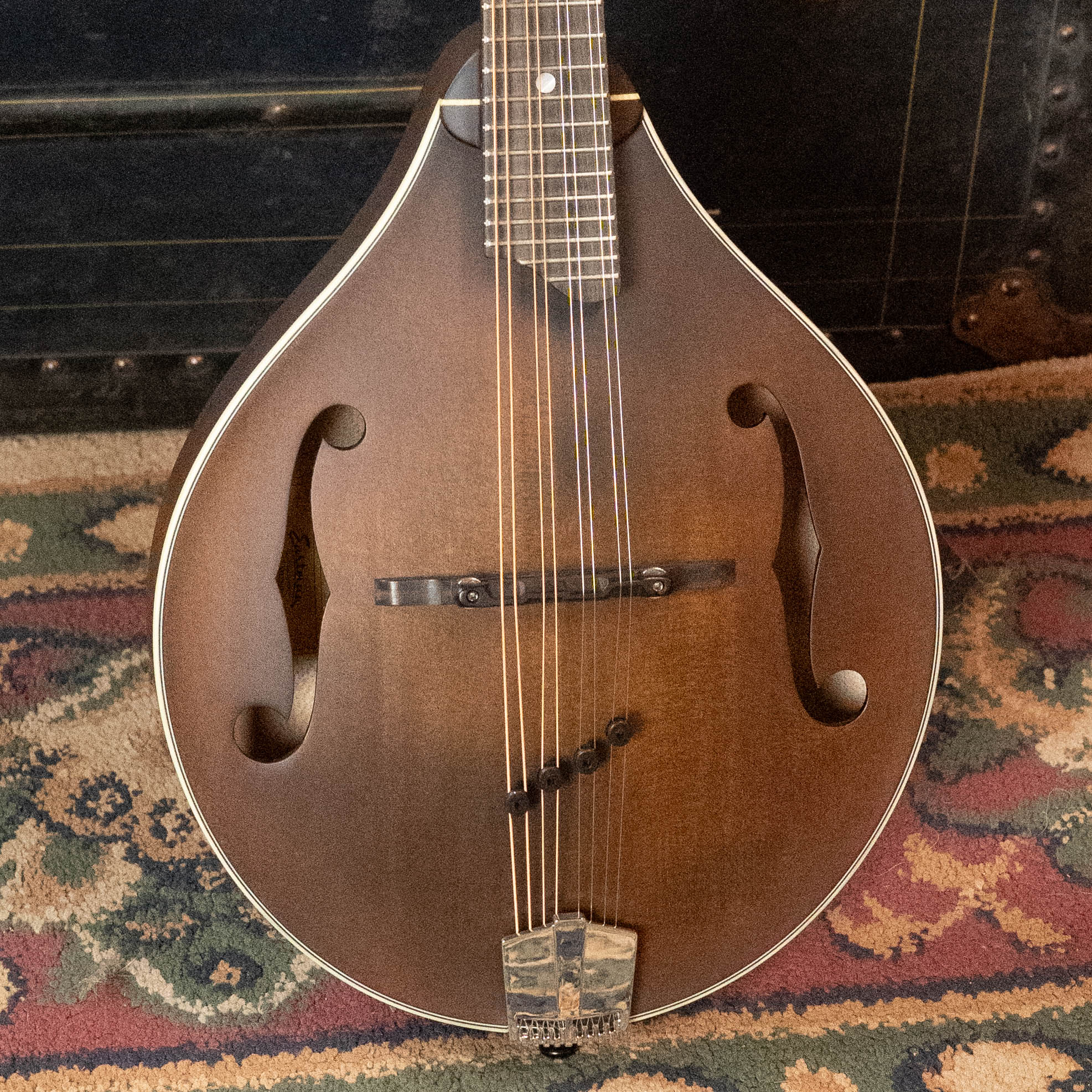 A close-up of an Eastman Guitars MD305 A-Style Hand-Carved Mandolin (#3596) with f-shaped sound holes and a solid spruce top, resting on a patterned rug with part of a black trunk visible in the background.