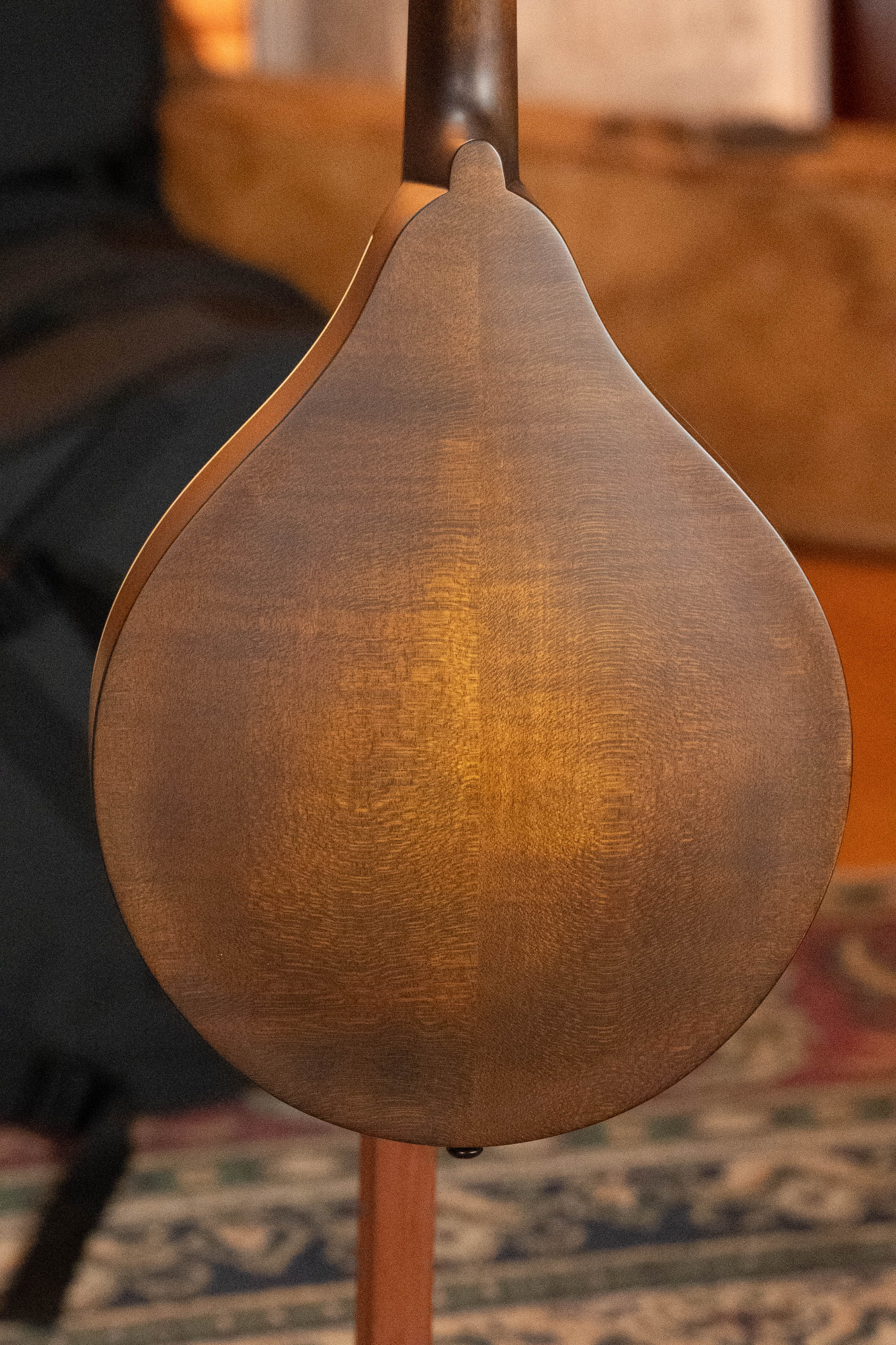 The image shows the back of an Eastman Guitars MD305 Spruce/Maple A-Style Hand-Carved Mandolin (#3596) with a rounded body and smooth brown finish, standing upright on a patterned rug.