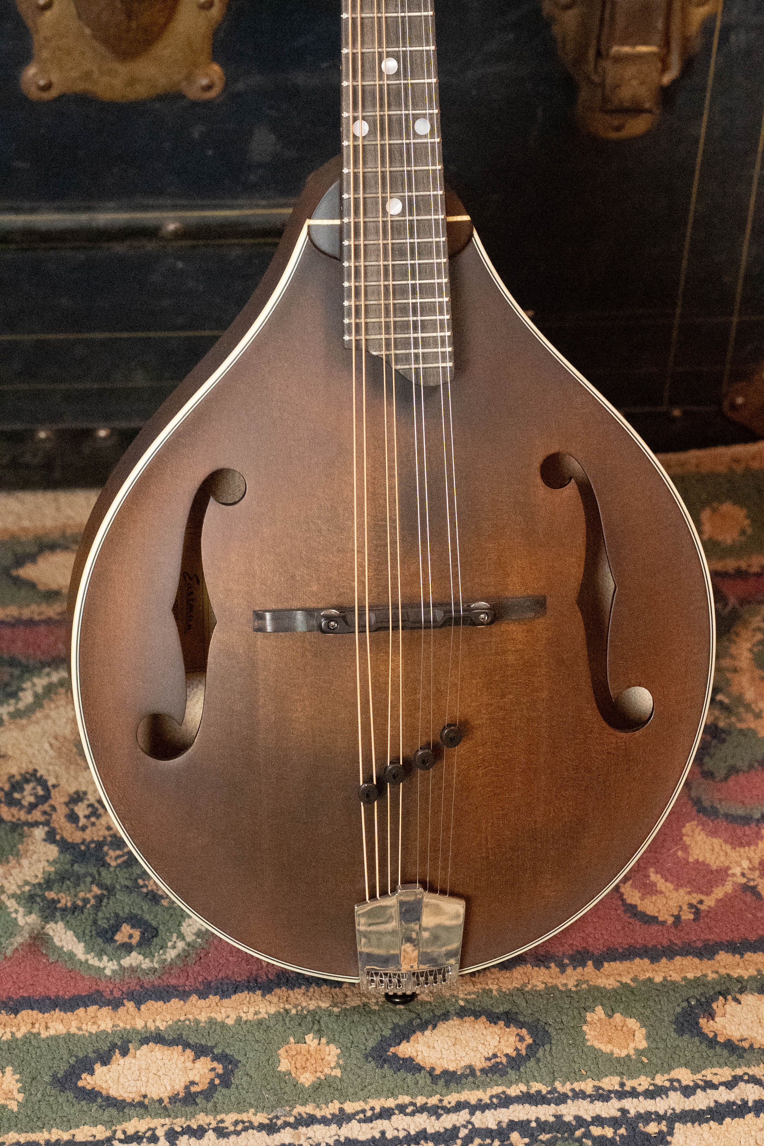 A close-up of the Eastman Guitars MD305 A-Style Hand-Carved Mandolin, featuring a solid spruce top, f-shaped sound holes, and sunburst finish, rests on a patterned rug with a vintage black trunk in the background.