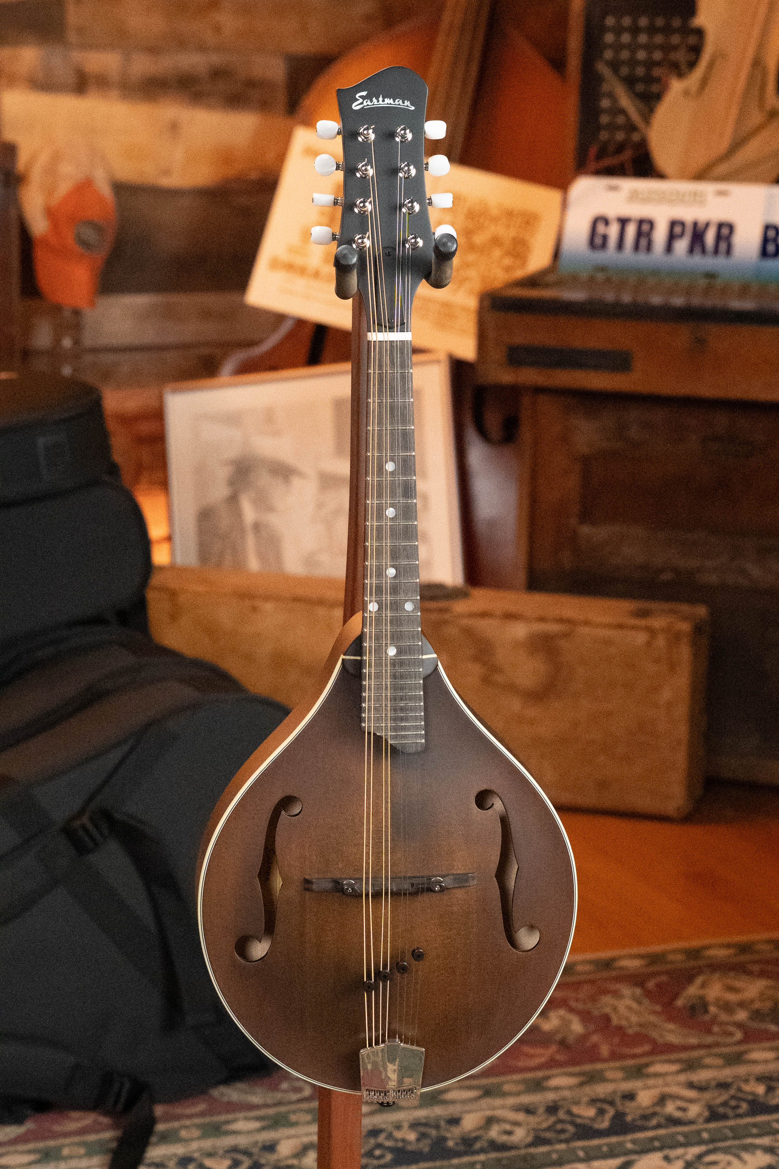 An Eastman Guitars MD305 Spruce/Maple A-Style Hand-Carved Mandolin #3596 with white tuning pegs stands upright indoors among music-themed decor, including framed photos, a guitar case, and a vintage-style license plate.