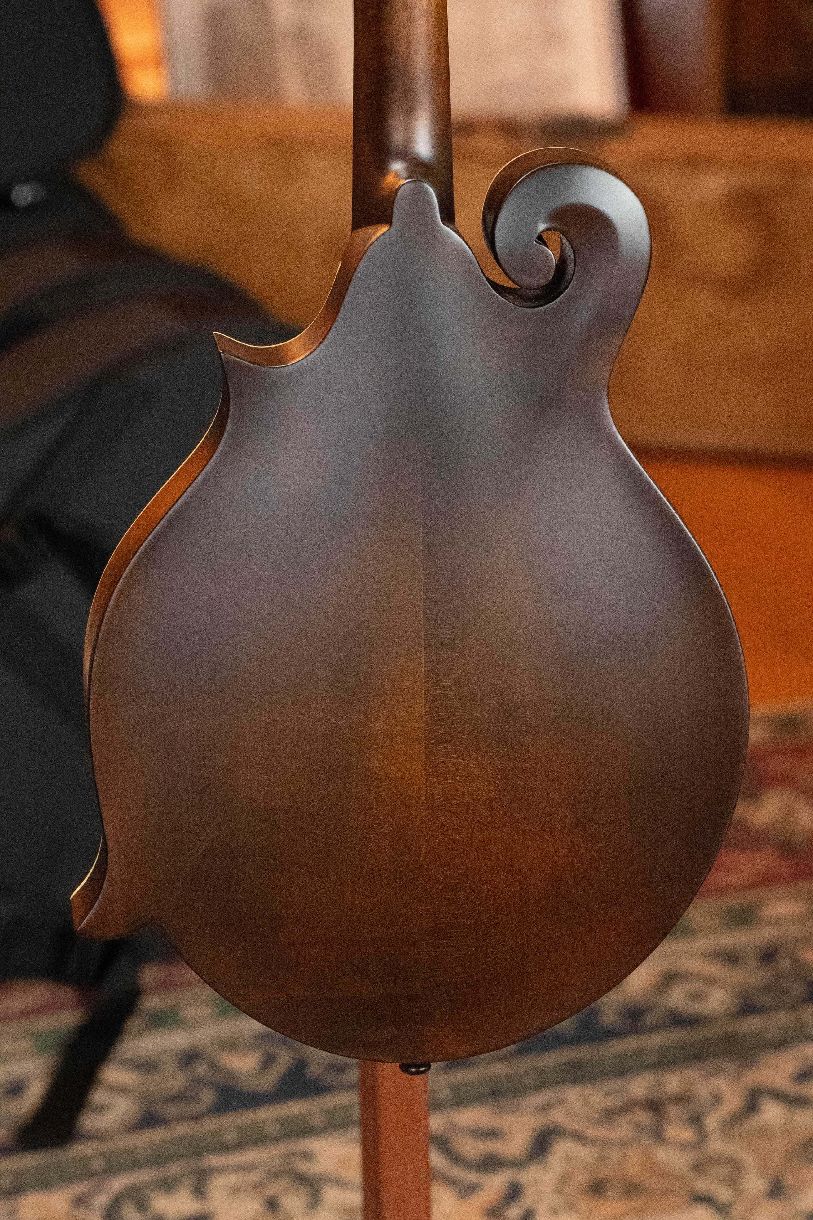 The image features the back of an Eastman Guitars MD315 F-Style Hand-Carved Mandolin (#0917) in a brown, glossy finish with a decorative scroll, displayed on a stand atop a patterned rug with blurred furniture and sheet music in the background.