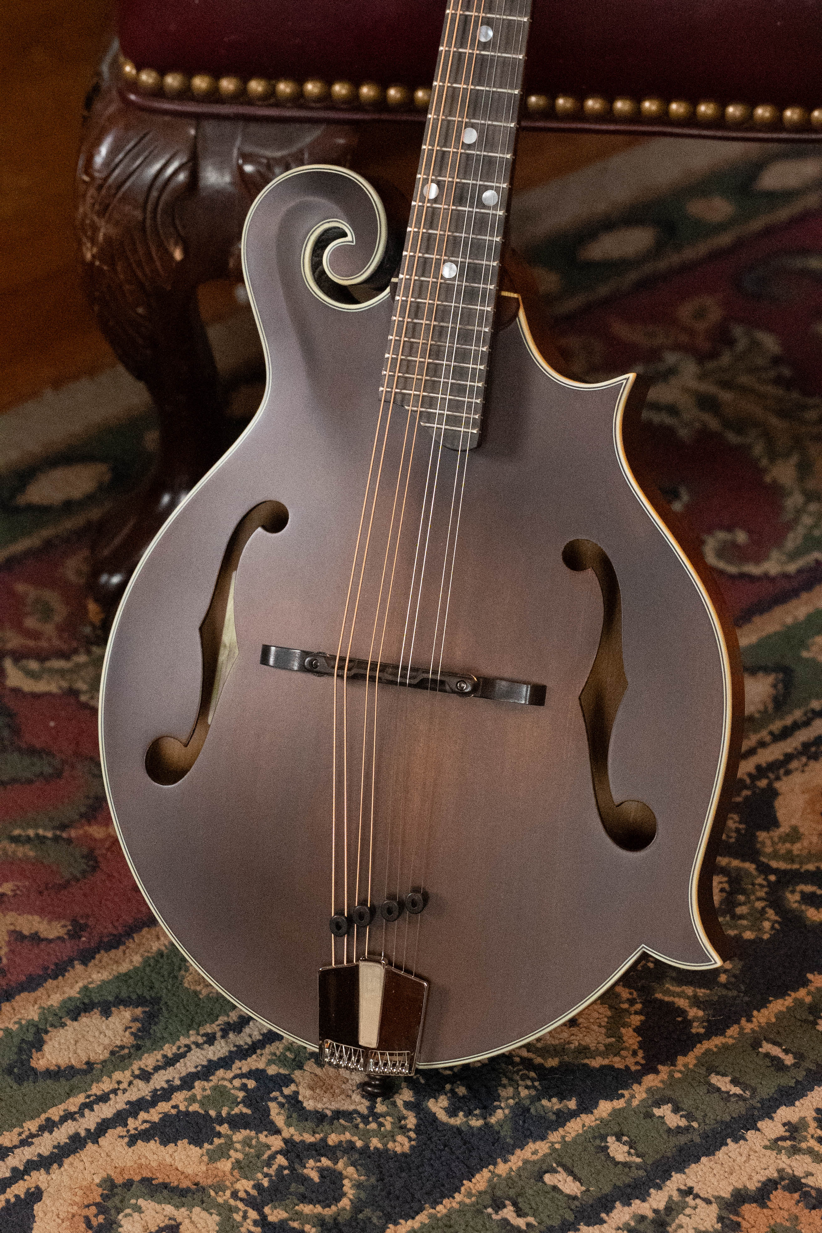 An Eastman Guitars MD315 F-Style Hand-Carved Mandolin (#0917), featuring a decorative scroll and f-shaped sound holes, stands on a patterned carpet beside a carved wooden chair.