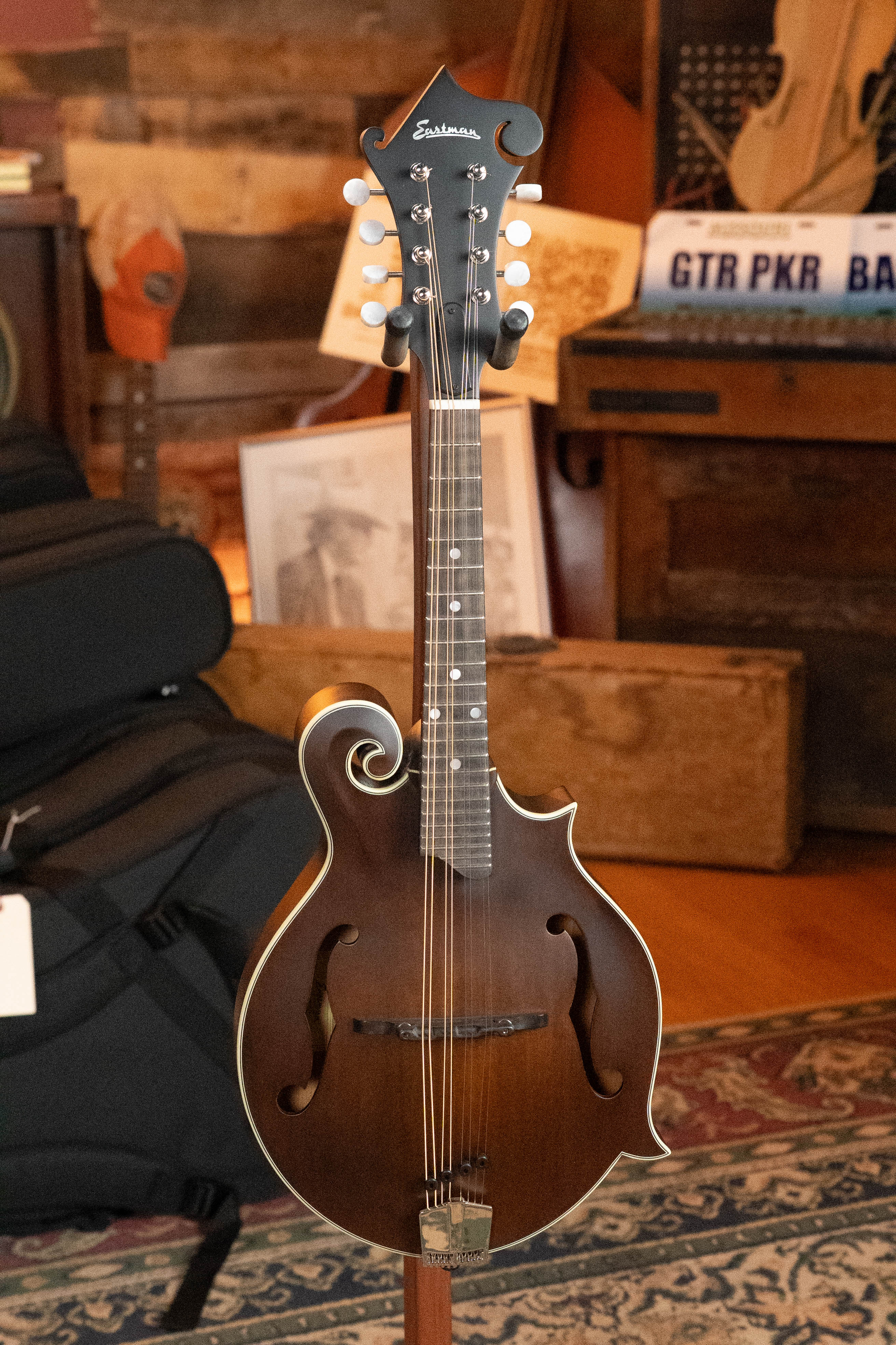 An Eastman Guitars MD315 F-Style Hand-Carved Mandolin (#0917) with a brown finish and decorative scroll headstock is displayed on a stand in a cozy room with wooden furniture, a patterned rug, and musical gear in the background.