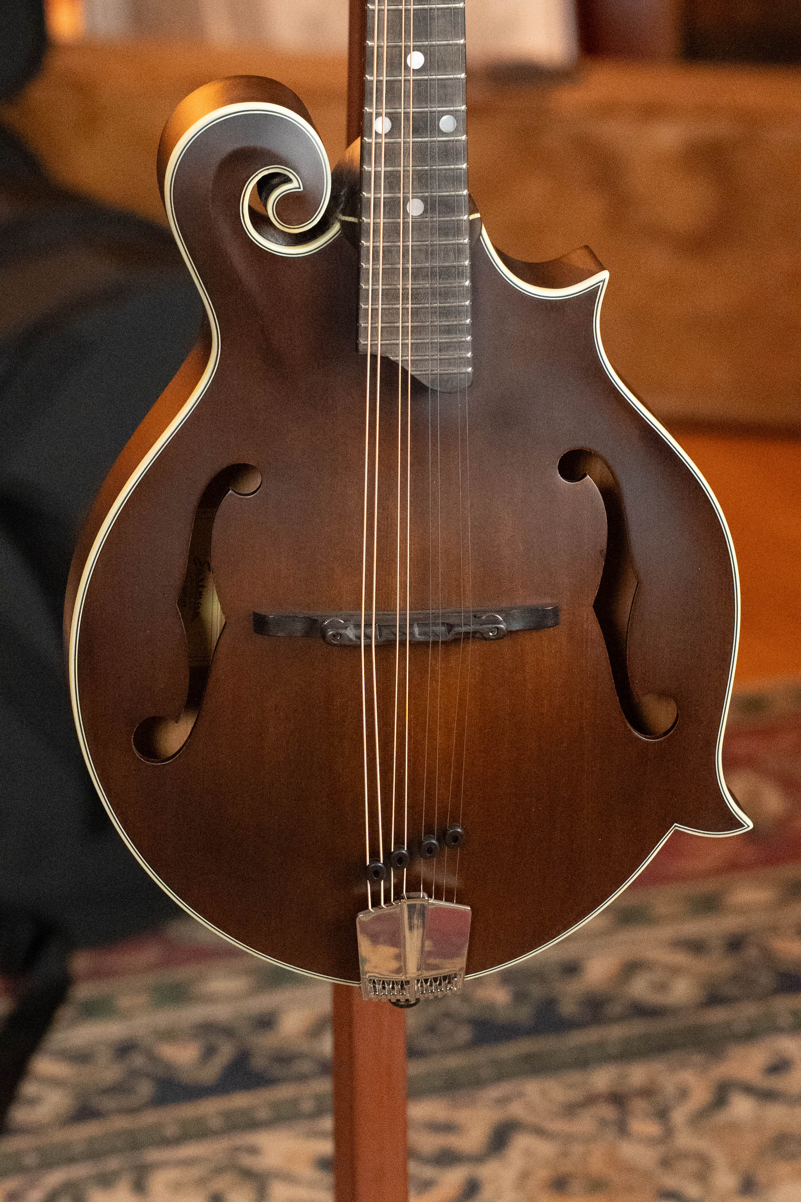 A close-up of the Eastman MD315 F-Style Hand-Carved Mandolin #0917 by Eastman Guitars, featuring a decorative scroll and f-holes, standing upright on a patterned rug with blurred furniture in the background.
