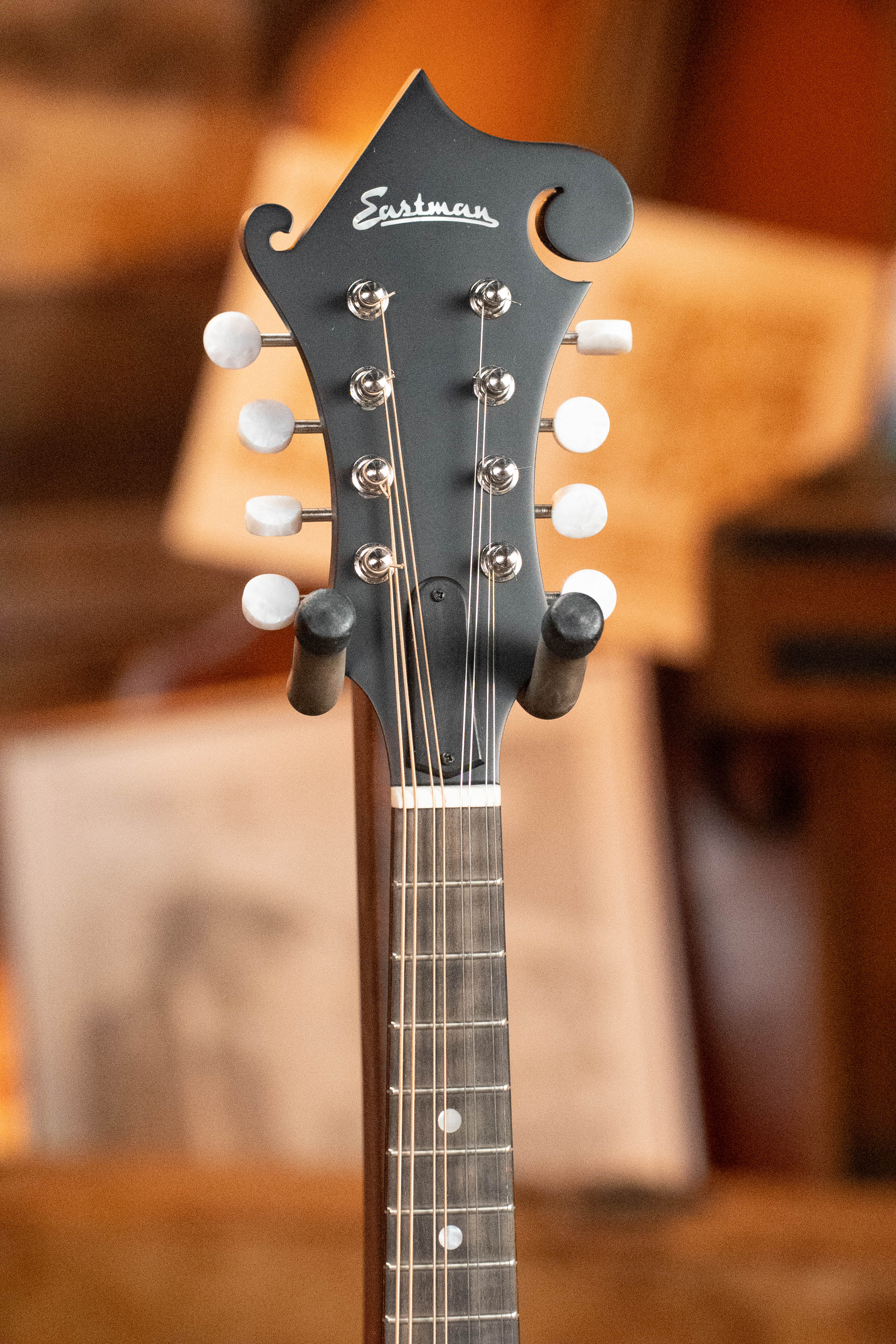 Close-up of the Eastman Guitars MD315 F-Style Hand-Carved Mandolin #0917 headstock, highlighting tuning pegs and strings against a blurred backdrop of sheet music and warm brown tones.