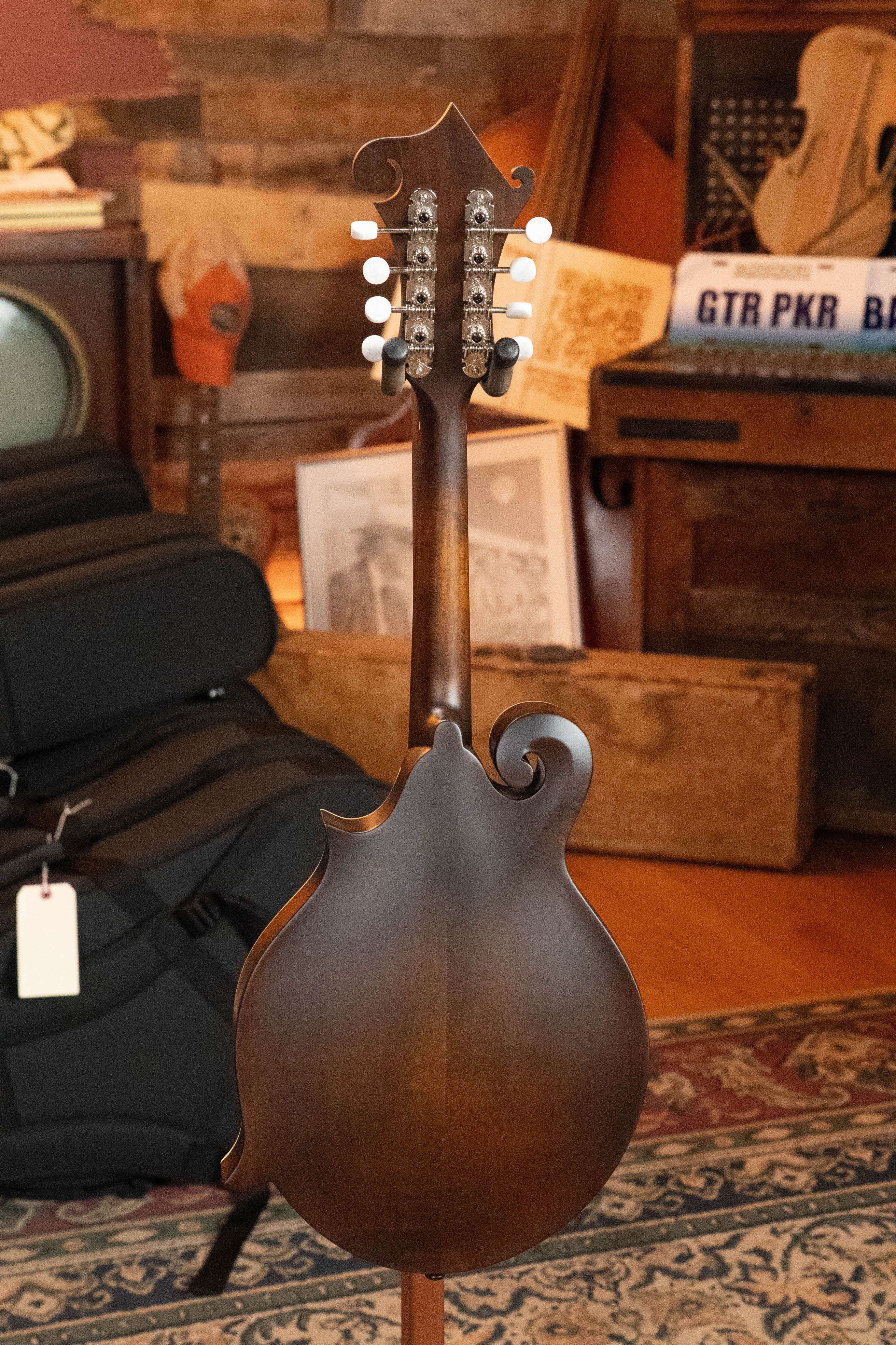 The Eastman Guitars MD315 F-Style Hand-Carved Mandolin #0917 stands upright, showing its back. Musical gear, framed photos, and rustic decor enhance the cozy room's atmosphere in the background.