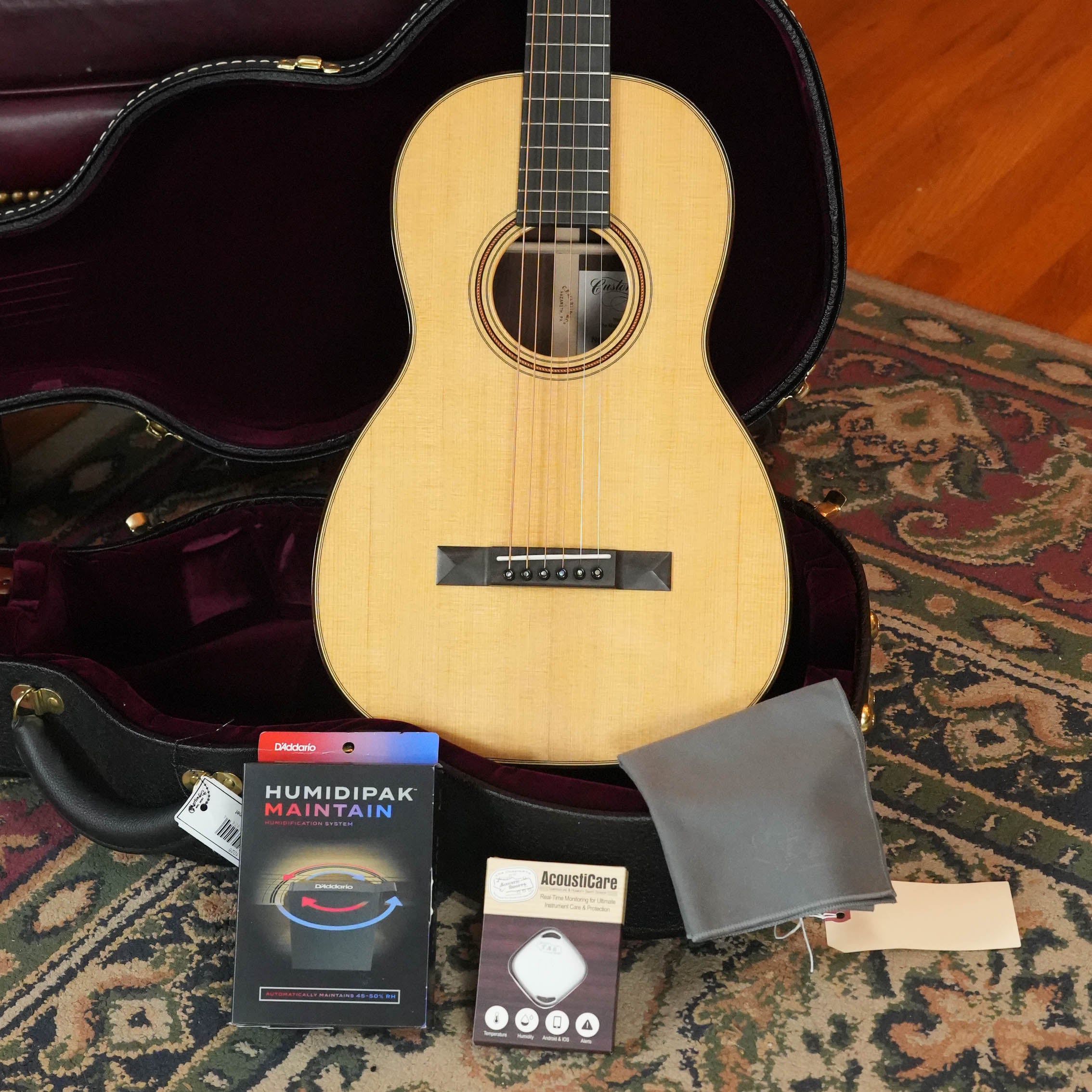A Martin & Co. Custom Shop Sitka/Rosewood Acoustic Guitar #6904 rests in an open case on a patterned rug, with accessories in front including a D’Addario Humidipak, Acousticare cleaner, and a gray cloth.