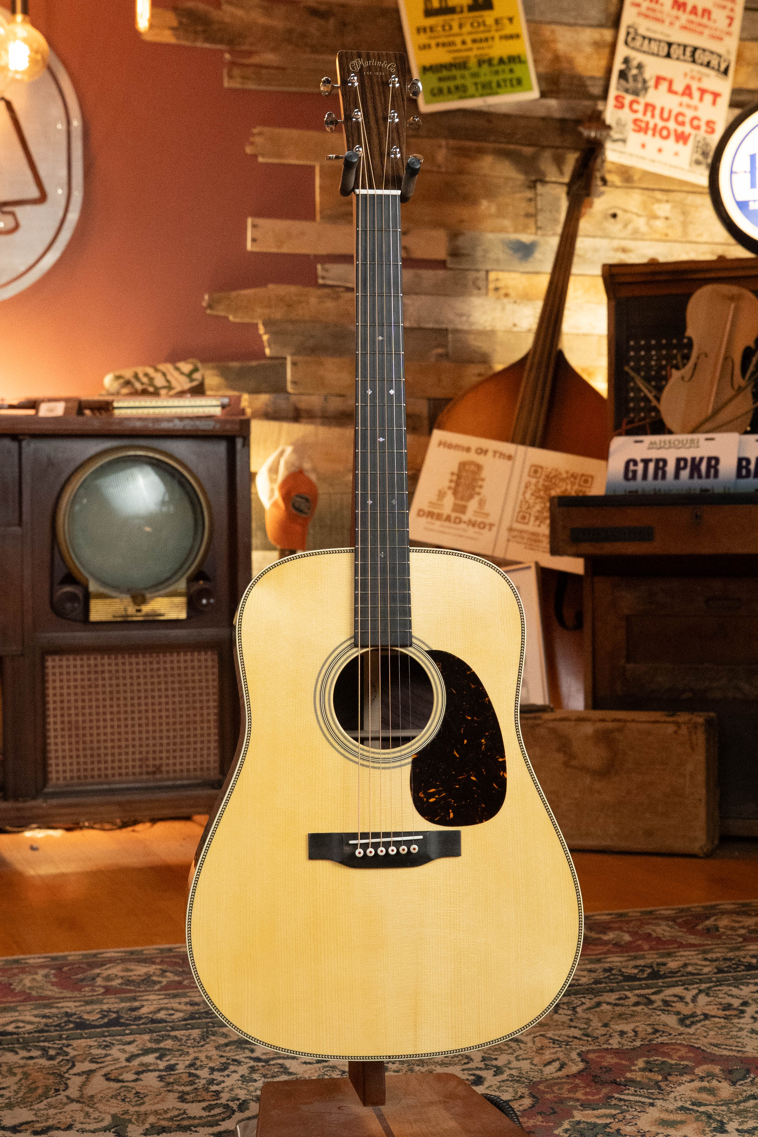 The Martin & Co. Custom Shop HD-28 Style Spruce/Wild Grain Rosewood Custom "D" Dreadnought Acoustic Guitar #7410 stands upright in a cozy vintage room with a retro TV, upright bass, and posters on wood-paneled walls.