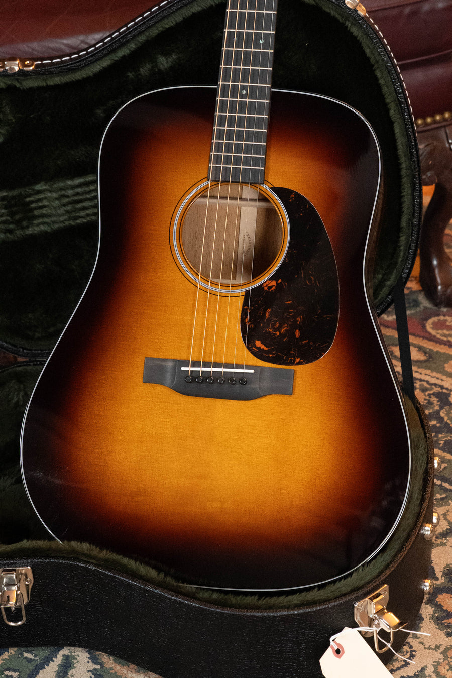 A Martin & Co. D-18 Spruce/Mahogany 1935 Gloss Sunburst Finish Dreadnought Acoustic Guitar (#5454) lies in an open, plush-lined black hard case with a small white tag, set on a patterned rug beside part of a leather chair.