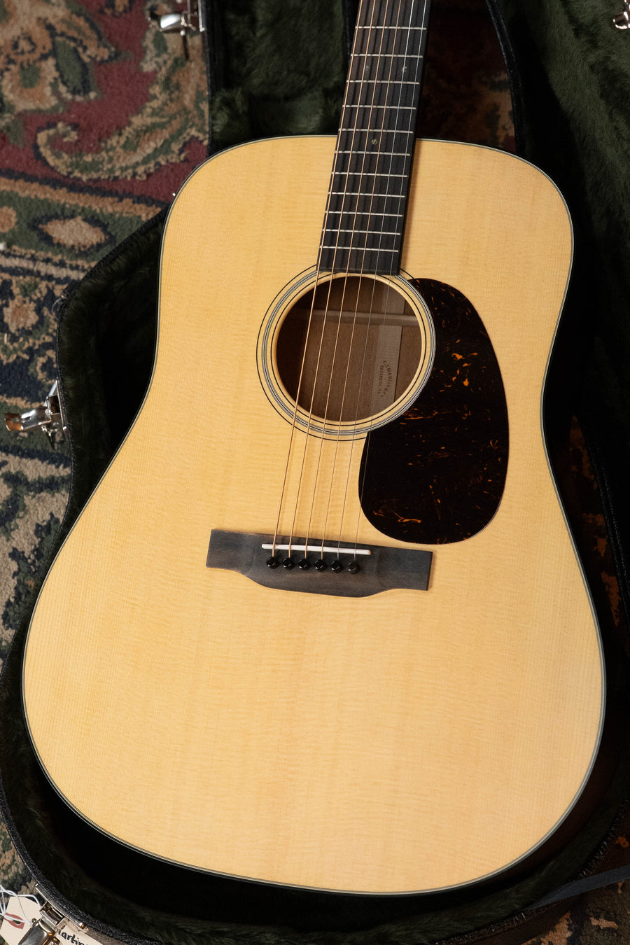 A close-up of a Martin & Co. D-18 Spruce/Mahogany Satin Finish Dreadnought Acoustic Guitar #1864, with a light spruce top, mahogany back and sides, dark pickguard, and six strings, resting in a black plush-lined hard case on patterned carpet.