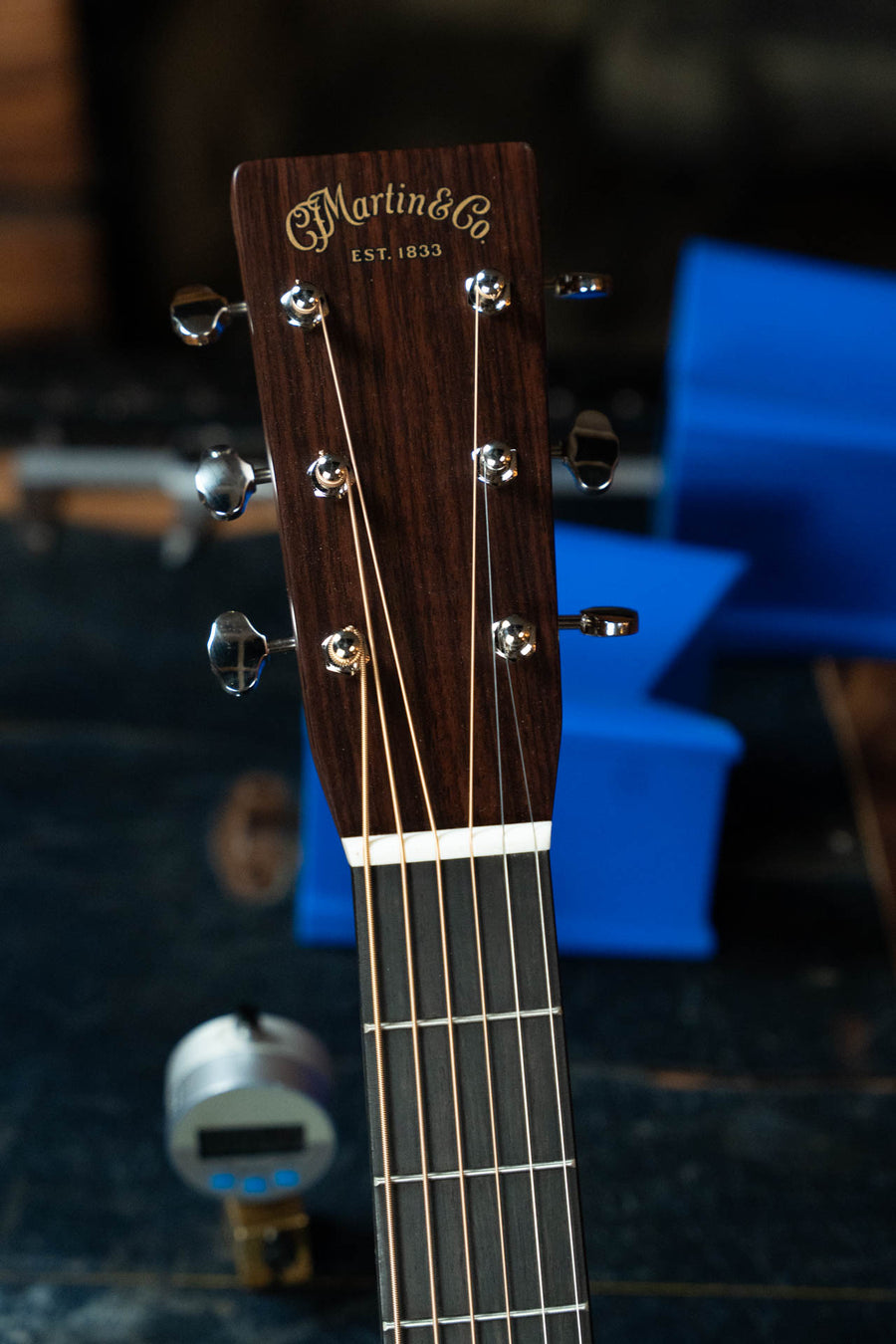 Close-up of a Martin & Co. D-28 Gloss Spruce/Rosewood Dreadnought Acoustic Guitar #1577 headstock, showing tuning pegs and strings with a blurred background featuring blue items and a small metallic device.