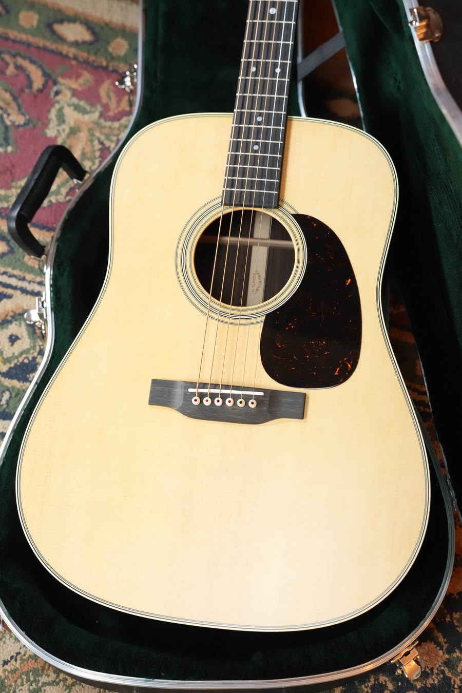 A Martin & Co. D-28 Satin Spruce/Rosewood Acoustic Guitar (#3679) with a dark pickguard rests in an open, velvet-lined case on a patterned rug.