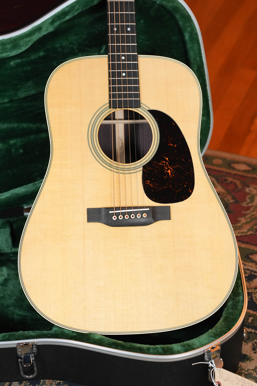 A Martin & Co. D-28 Satin Spruce/Rosewood Acoustic Guitar #7987 with a tortoiseshell pickguard is displayed in a green velvet-lined hard case atop a patterned rug. This six-string features a light spruce top and timeless design.