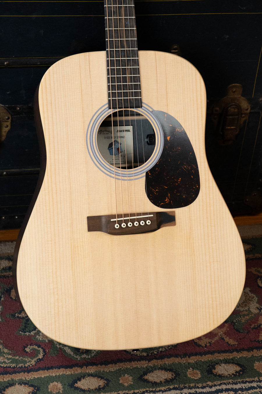 A Martin & Co. D-X2E Billy Strings Spruce/HPL Rosewood Dreadnought Guitar with six strings and black pickguard stands upright on a patterned rug before a dark vintage-style chest.
