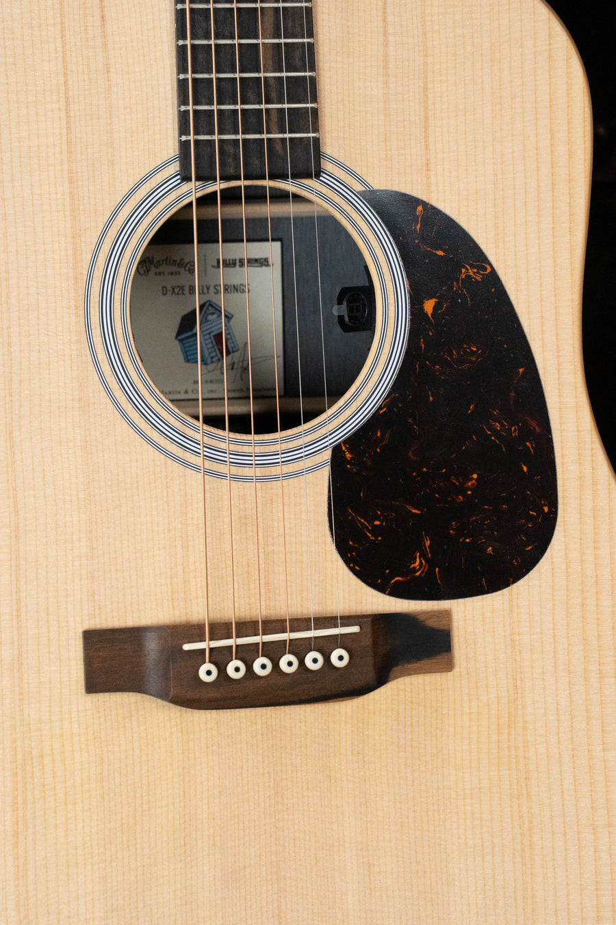 Close-up of a Martin & Co. D-X2E Billy Strings Spruce/HPL Rosewood dreadnought guitar body, highlighting its spruce top, soundhole, bridge, and strings with a light finish and dark tortoiseshell-patterned pickguard.