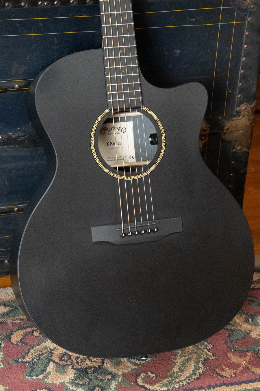 The Martin & Co. GPC-X1E All Black HPL Grand Performance Cutaway Guitar with Martin E1 Electronics (#1604) leans against a vintage trunk on a patterned rug, its logo visible inside the sound hole.