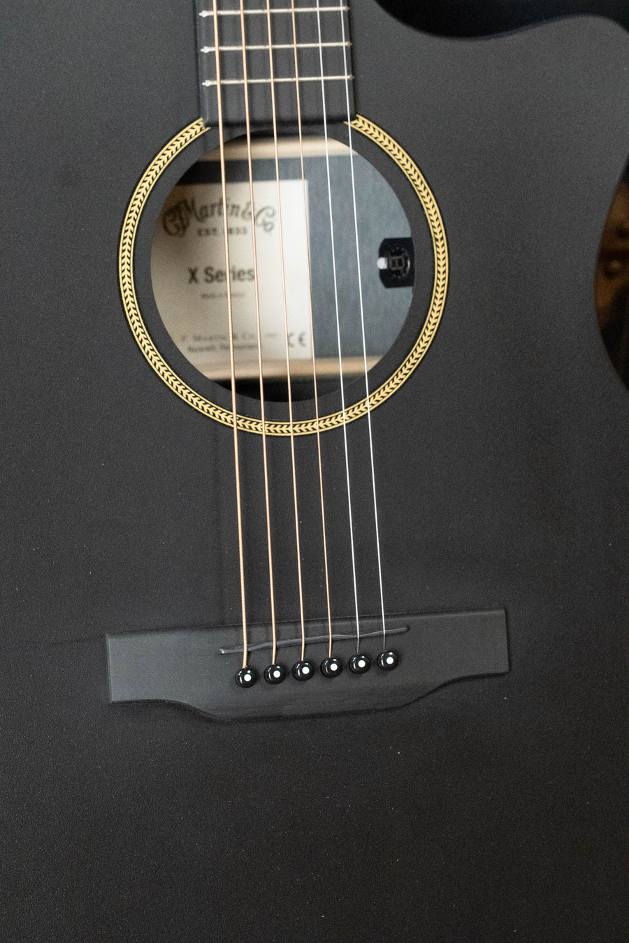 Close-up of a Martin & Co. GPC-X1E All Black HPL Grand Performance Cutaway Guitar with E1 Electronics, showing strings, bridge, and soundhole with decorative ring. The inside label reads Martin & Co. X Series.