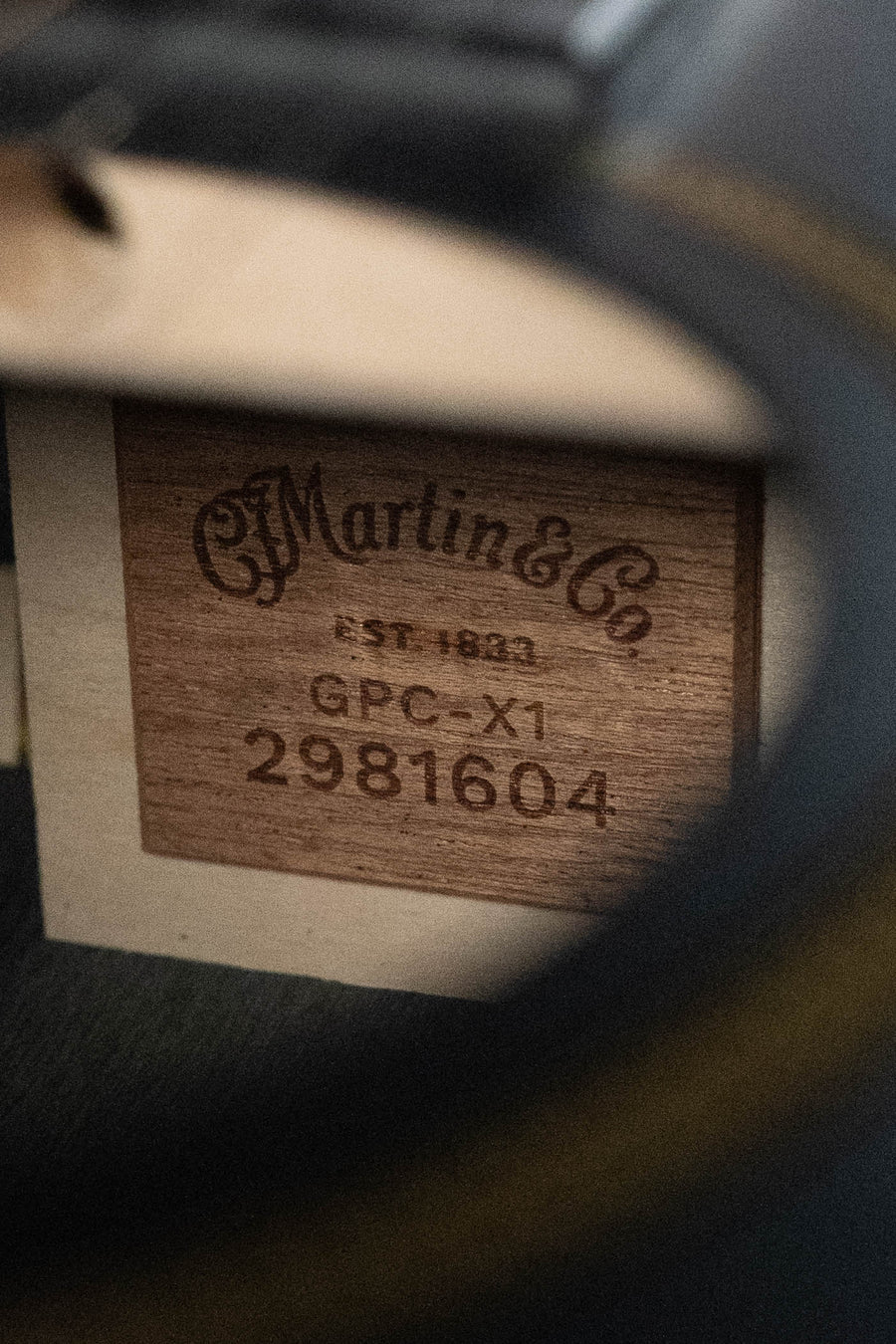 Close-up of a wooden label inside a Martin & Co. GPC-X1E All Black HPL Grand Performance Cutaway Guitar w/Martin E1 Electronics #1604, displaying the logo, EST. 1833, model info, and serial number.