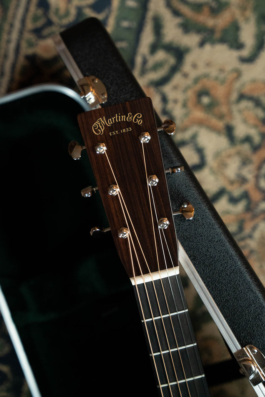Close-up of a Martin & Co. HD-28 1935 Sunburst Spruce/East Indian Rosewood Dreadnought Acoustic Guitar (#5446) headstock, resting on its open black hard case with a patterned carpet beneath.