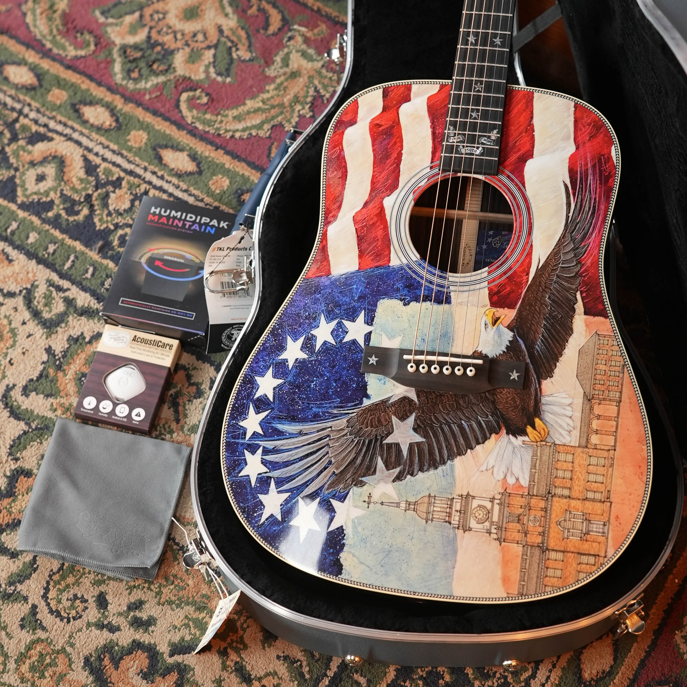 An open case displays the Martin & Co. HD-28 Semiquincentennial Limited Edition Acoustic Guitar #7873—featuring a custom American flag, bald eagle, and historic building design—plus humidipak, tuner, cloth, and strings on a patterned rug.