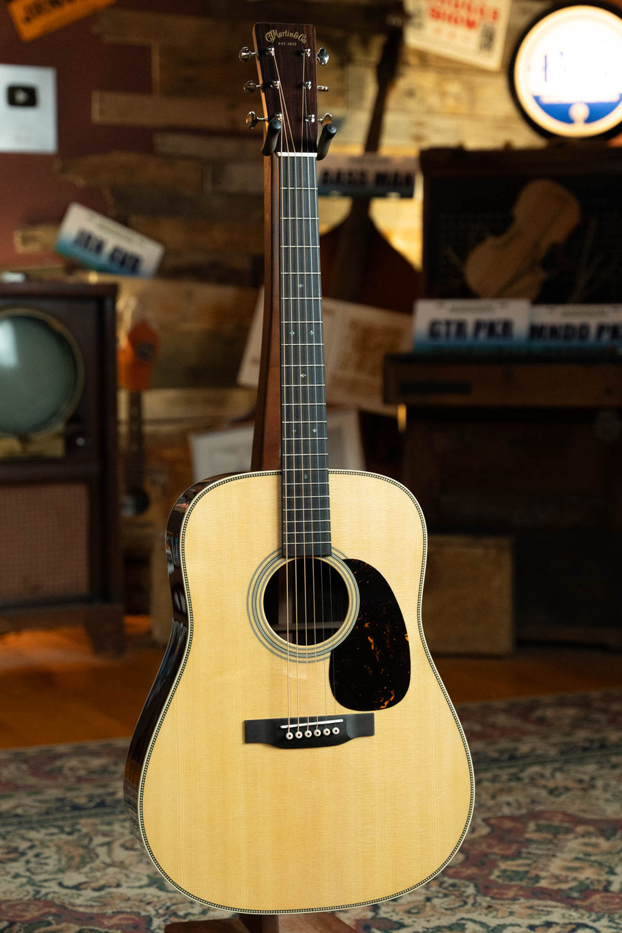 The Martin & Co. HD-28 Spruce/East Indian Rosewood Dreadnought Acoustic Guitar #8420 stands upright on a patterned rug in a cozy, vintage room with wood paneling, an old TV, and assorted signs in the background.