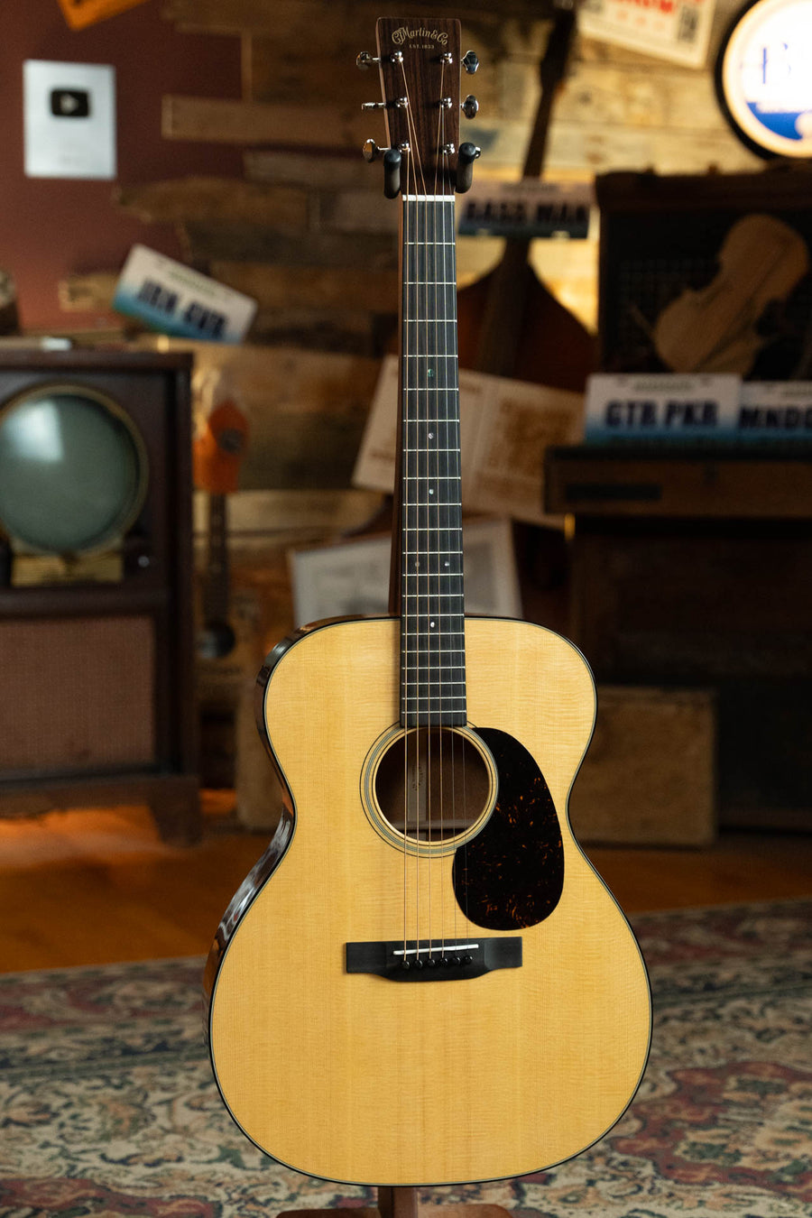 A Martin & Co. OOO-18 Spruce/Mahogany OOO Acoustic Guitar #8901 stands on a patterned rug in a cozy, vintage-style room featuring wood paneling and retro decor.