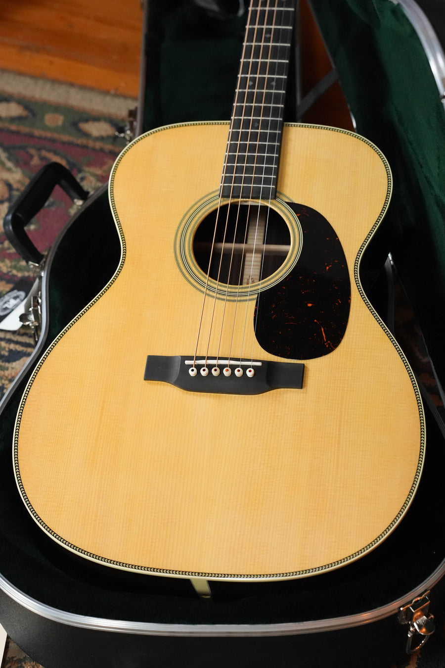 A Martin & Co. OOO-28 Gloss Spruce/Rosewood acoustic guitar (#7960) with a light wood top and black pickguard rests in an open hard case, while part of a patterned rug is visible in the background.