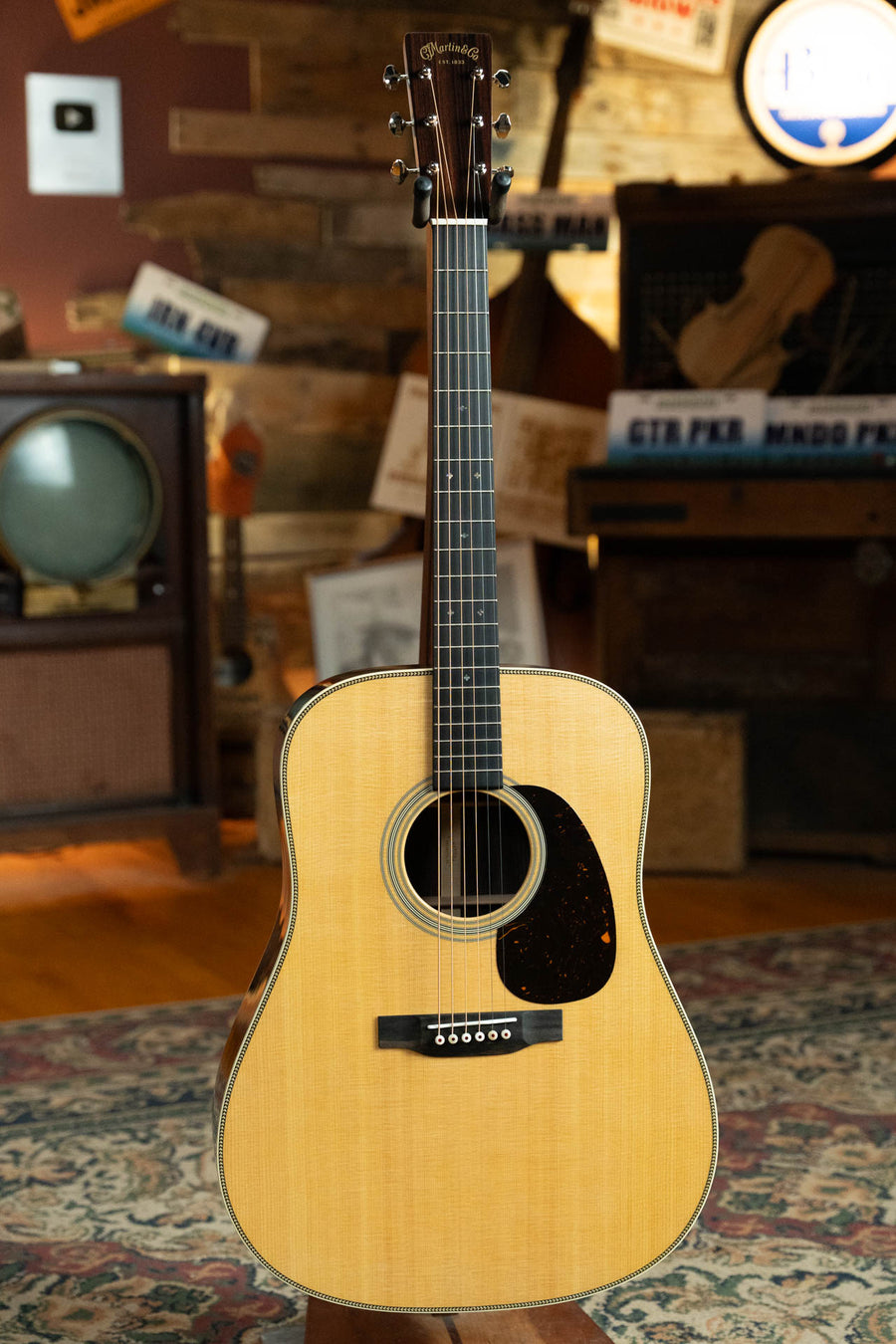 A Martin & Co. Super HD-28 Spruce/Indian Rosewood Dreadnought Acoustic Guitar #8394 stands upright on a patterned rug in a cozy vintage room with wood walls, a retro TV, and music-themed decor in the background.
