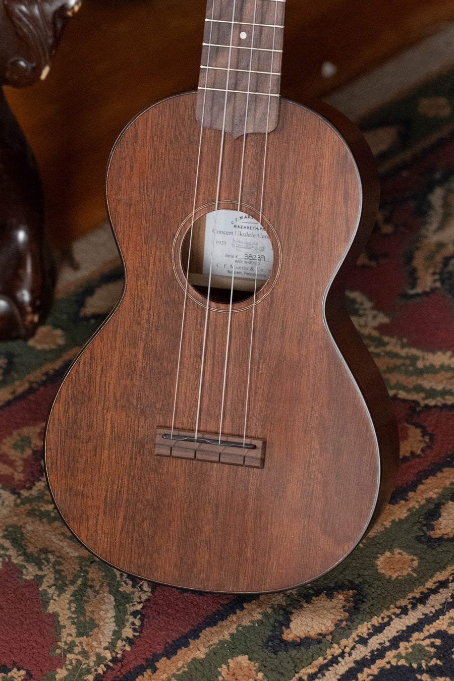 A close-up of the Martin & Co. Centennial Concert All Mahogany Ukulele #8239 stands upright on a patterned carpet, highlighting its strings, sound hole, and part of the neck.