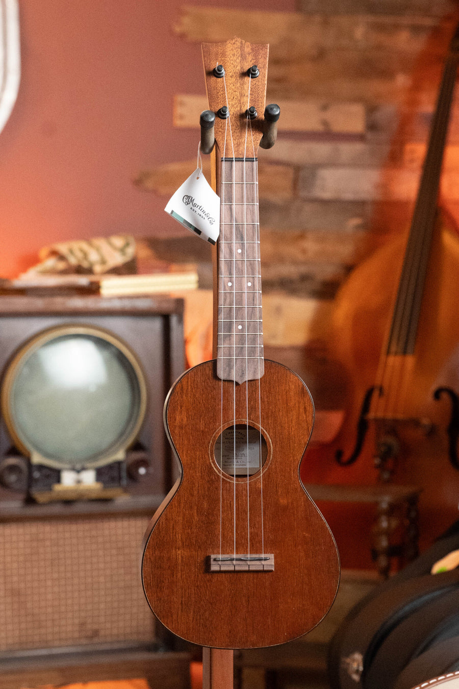 A Martin & Co. Centennial Concert All Mahogany Ukulele #8239 with a tag is on display in front of a vintage TV and radio, with part of a stand-up bass visible in the background.