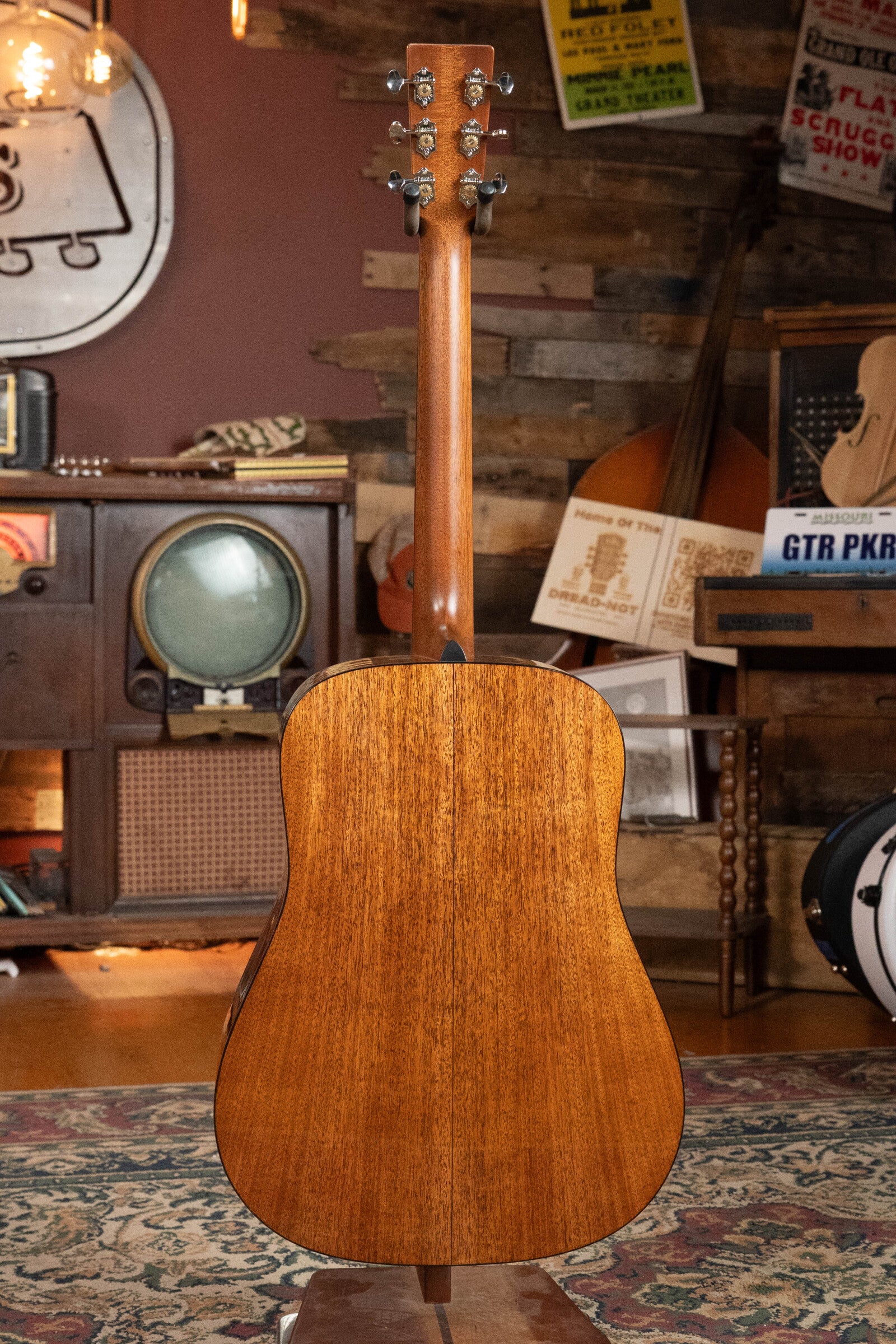 The Martin & Co. Custom Shop D-18V Adirondack/Mahogany Dreadnought Acoustic Guitar #6905 is displayed from the back, its rich brown grain visible as it stands upright in a cozy, retro room filled with musical instruments and nostalgic decor.