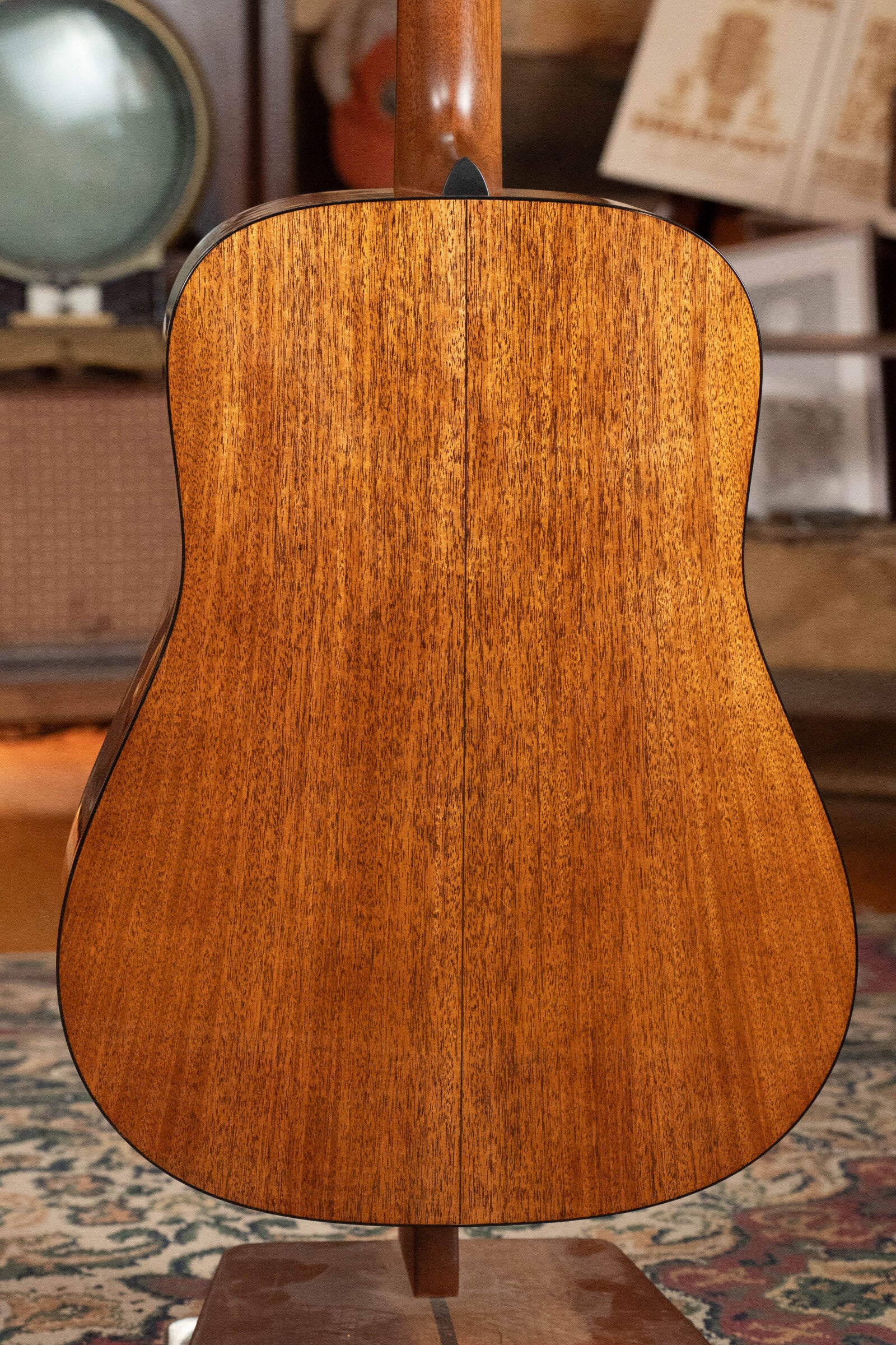 The back of a Martin & Co. Custom Shop D-18V Adirondack/Mahogany Dreadnought Acoustic Guitar #6905, featuring polished mahogany and fine grain, rests on a stand, its rich brown tone complementing the vintage decor and patterned rug behind it.