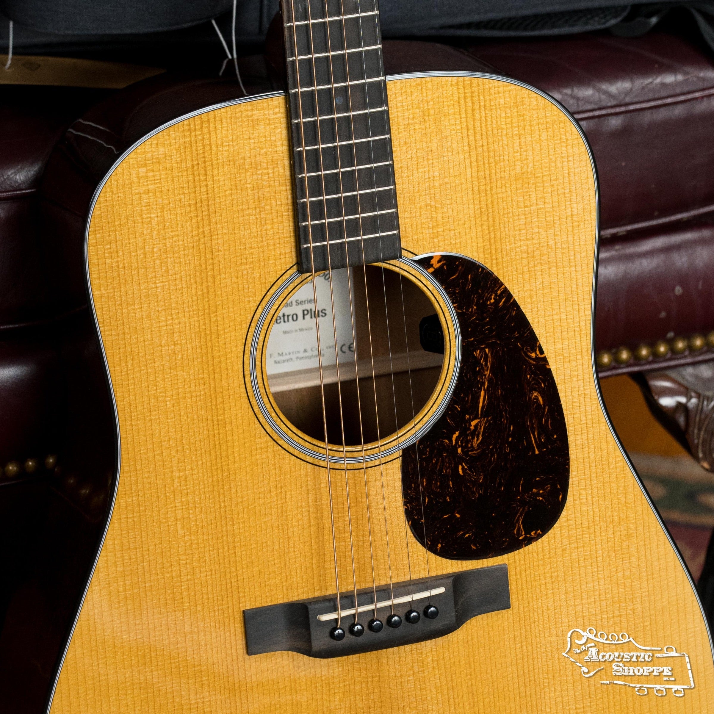 A close-up of the Martin & Co. DE Retro Plus Torrefied Spruce/Mahogany Dreadnought #0808 guitar shows its natural wood finish, tortoiseshell pickguard, and soundhole label as it rests on a leather seat, promising vintage-inspired tone.