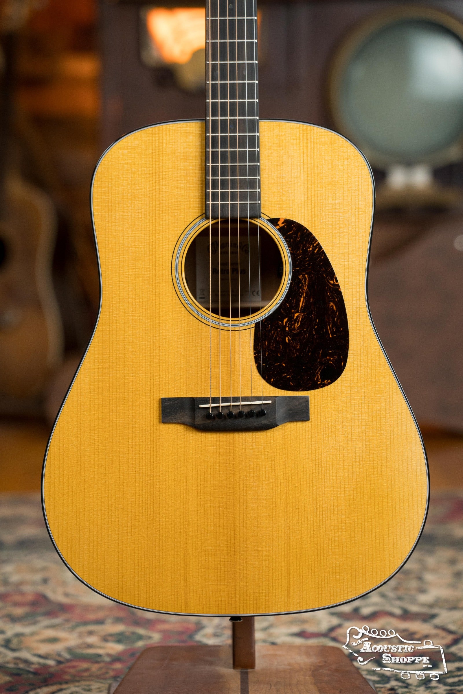 A close-up of the Martin & Co. DE Retro Plus Torrefied Spruce/Mahogany dreadnought guitar (#0808) showcases its wooden body, black pickguard, and strings against a blurred background with musical instruments and a TV.