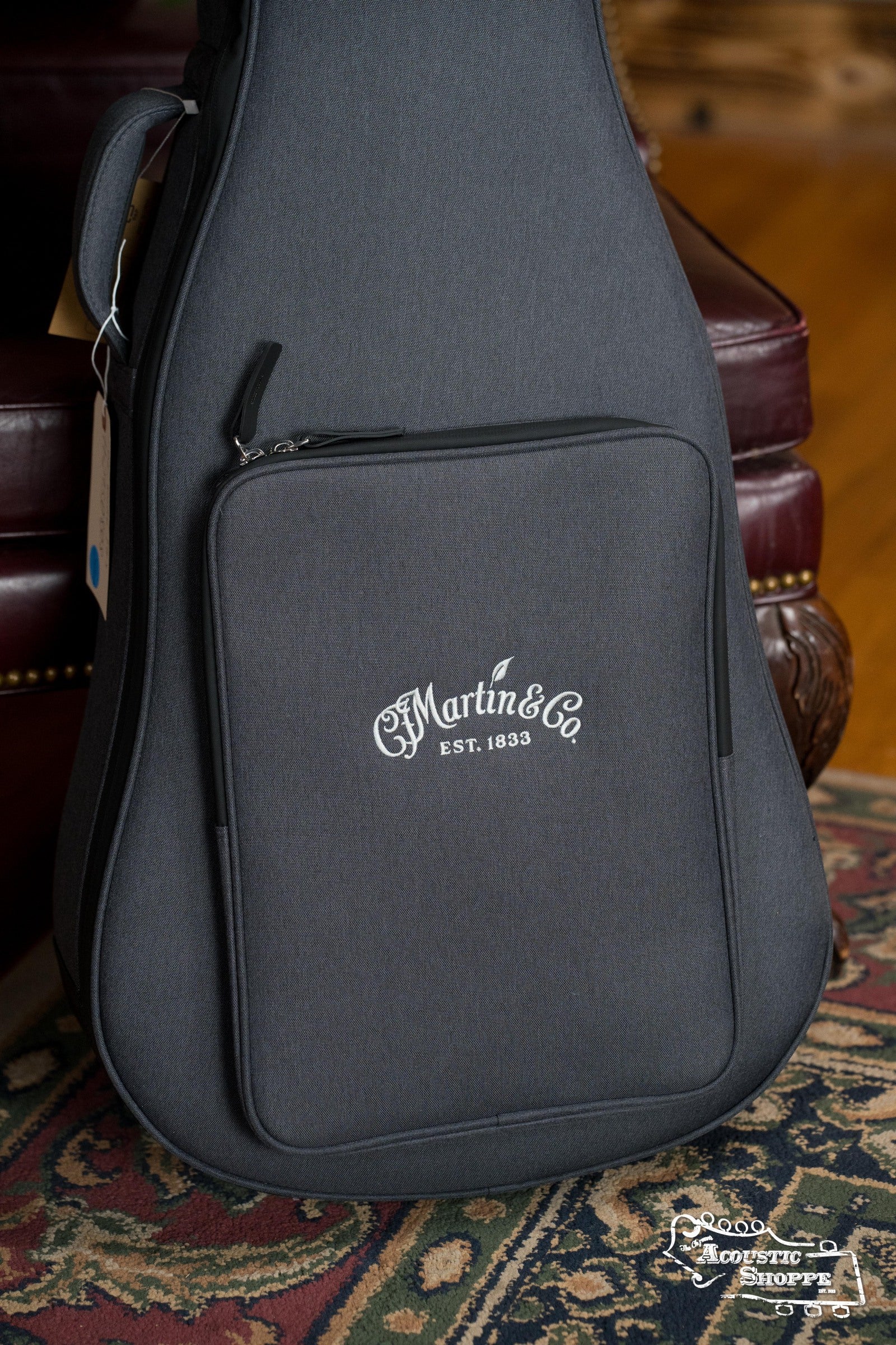 A gray Martin & Co. case for the DE Retro Plus Torrefied Spruce/Mahogany Dreadnought stands upright on a patterned rug by a leather chair, featuring the classic logo and Est. 1833 that reflect its vintage-inspired style.