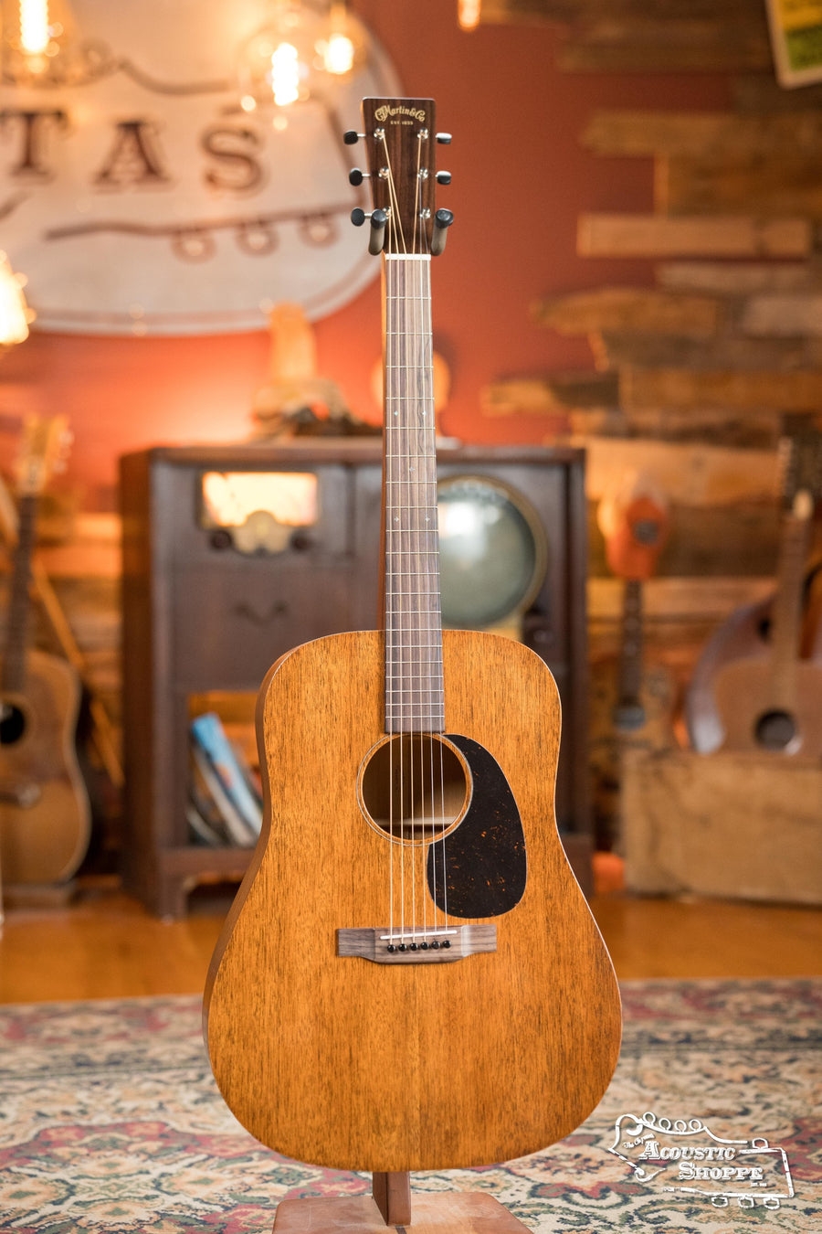 The Martin & Co. D-15M All Mahogany Dreadnought Acoustic Guitar #1493 stands upright on a patterned rug in a vintage-inspired room filled with musical instruments and wooden furniture, showcasing its natural wood finish.