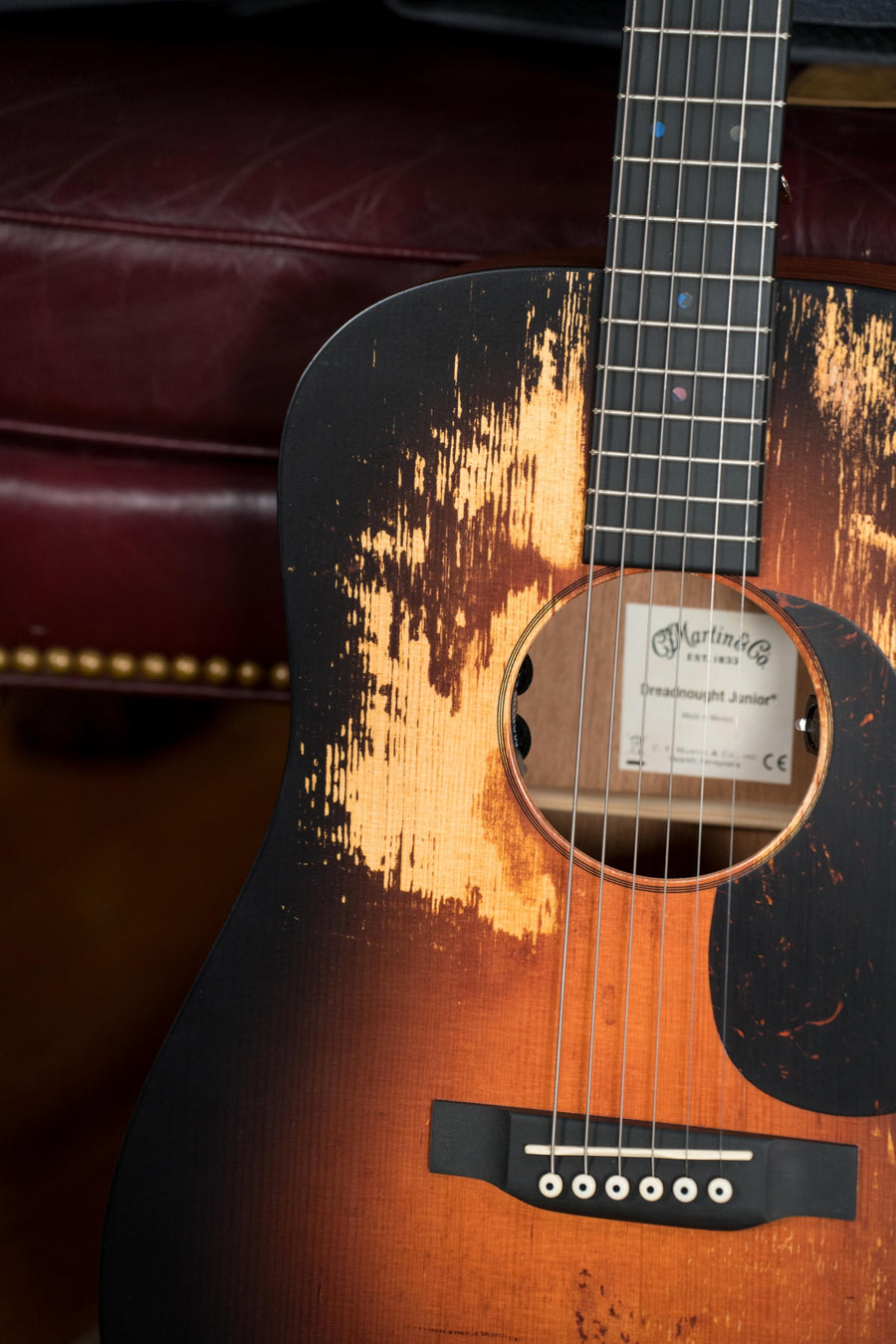 A close-up of the Martin & Co. D Jr E Streetlegend Burst Spruce/Sapele Aged-Look Dreadnought Guitar w/Martin Electronics #2363 shows its distressed vintage finish and exposed wood as it rests on a red leather seat with brass studs.