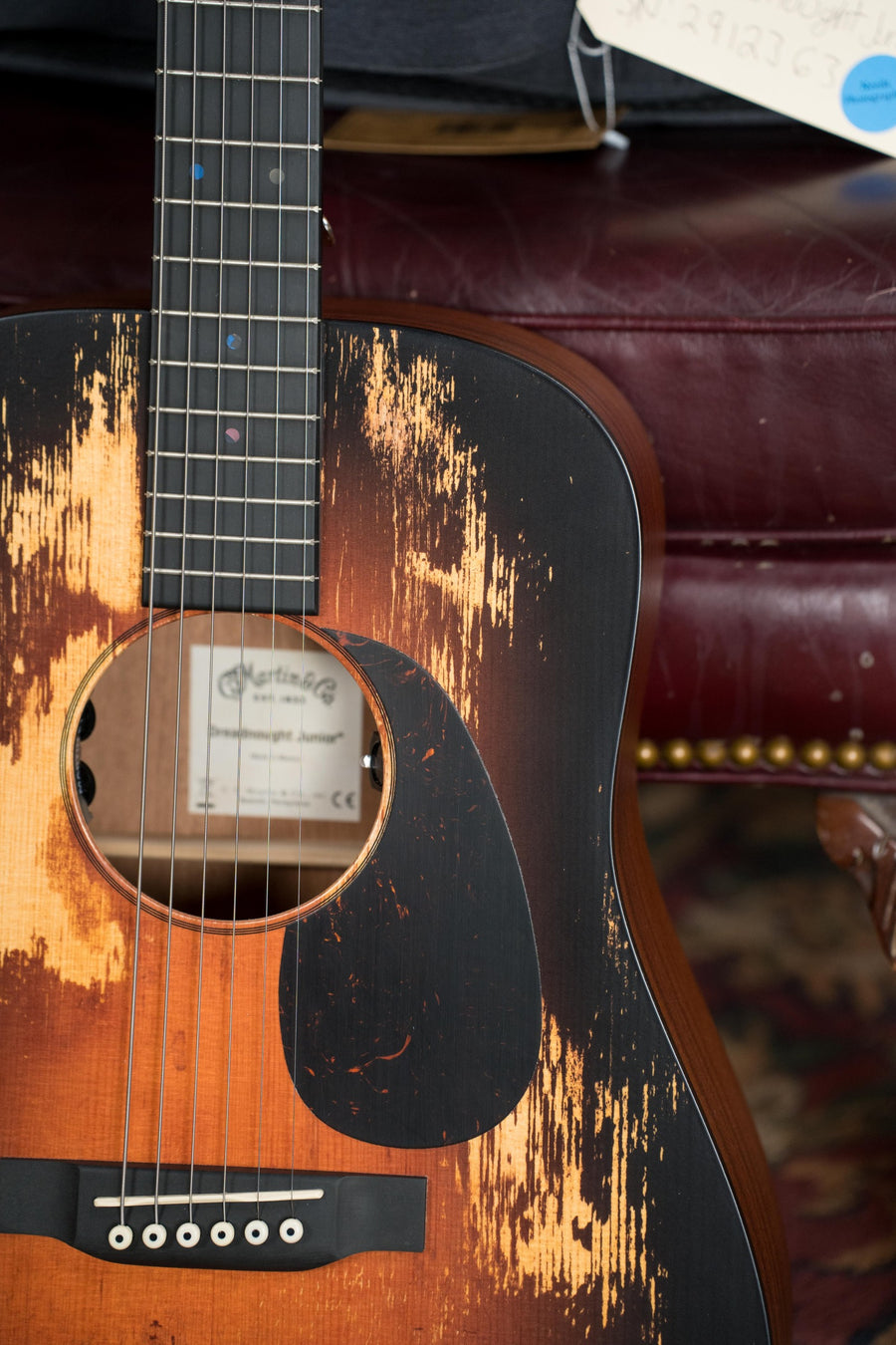 Close-up of a Martin & Co. D Jr E StreetLegend Burst Spruce/Sapele Aged-Look Dreadnought Guitar w/Martin Electronics #2363, featuring a vintage, worn finish and resting on a red tufted leather couch with brass studs.