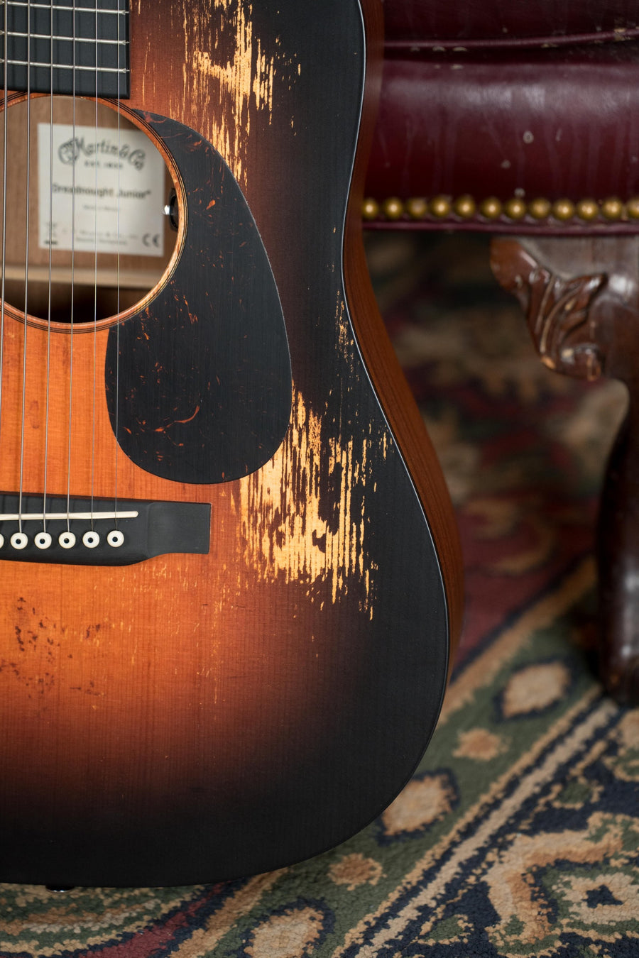 A worn Martin & Co. D Jr E Streetlegend Burst Spruce/Sapele Aged-Look Dreadnought Guitar w/Martin Electronics (#2363) shows scratches and fading beside a carved chair leg on a patterned rug.