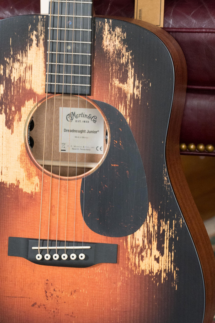 A close-up of the *New 2025 Model* Martin & Co. D Jr E Streetlegend Burst, a compact dreadnought guitar with aged spruce/sapele finish, black pickguard, and visible strings, resting on a red tufted background.