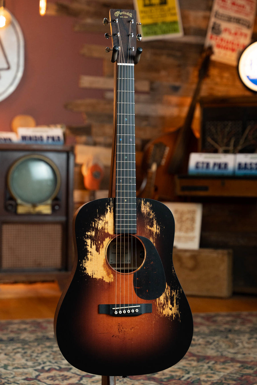 A Martin & Co. D Jr E Streetlegend Burst Spruce/Sapele Aged-Look Dreadnought Guitar w/Martin Electronics (#3872) with a distressed finish stands upright in a cozy room adorned with wood-paneled walls, vintage TV, posters, and music gear.