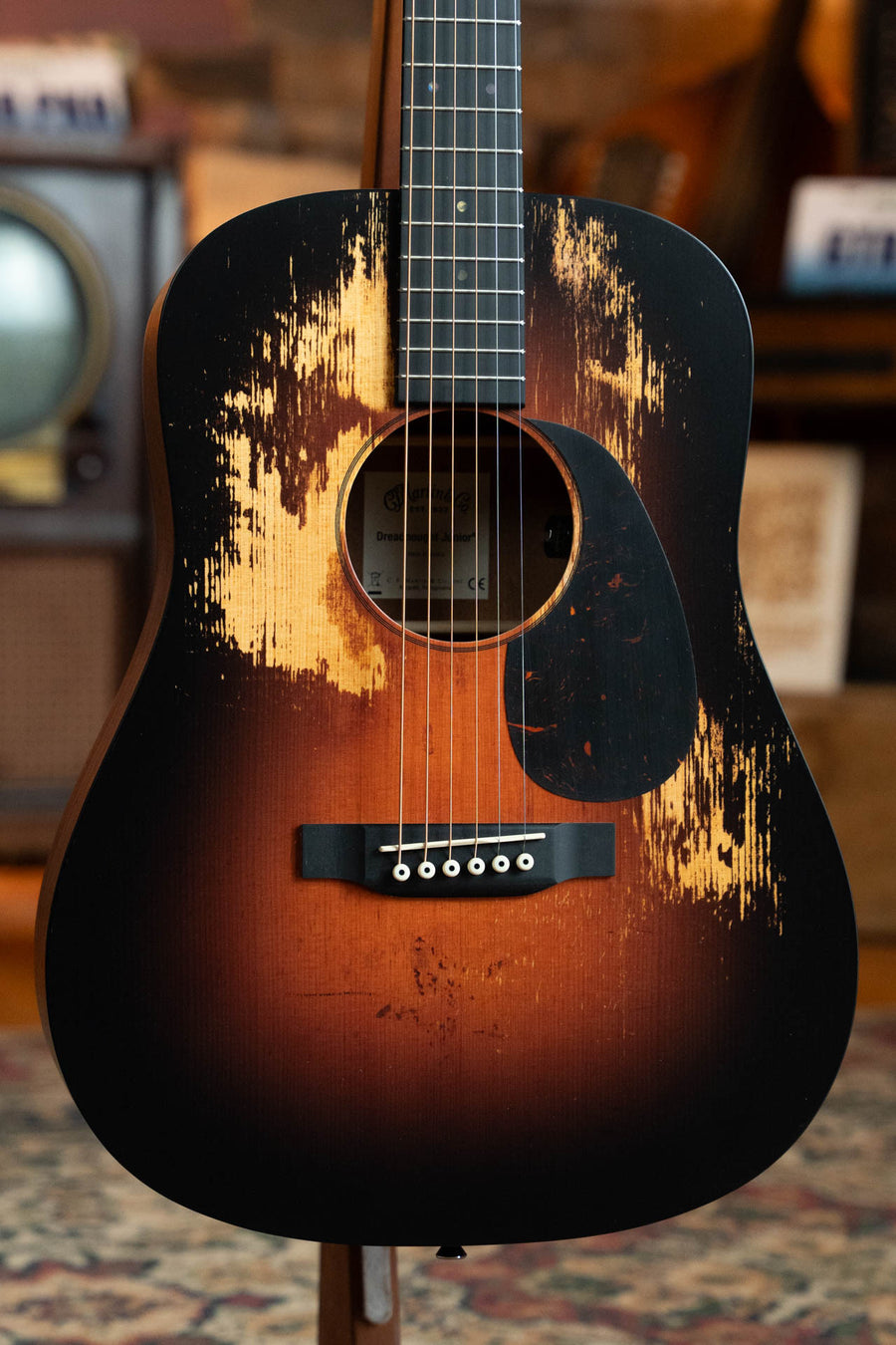 A close-up of the Martin & Co. D Jr E Streetlegend Burst Spruce/Sapele Aged-Look Dreadnought Guitar with exposed wood, black pickguard, and vintage-style backdrop. Features built-in Martin Electronics. Model #3872.